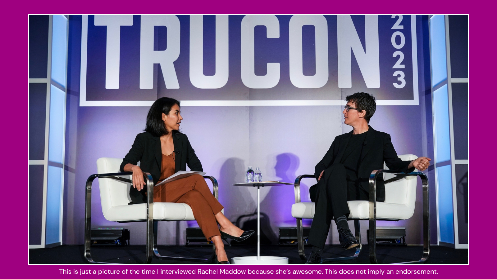 Two women seated on stage in an interview setting at the TechCrunch 2023 conference. One woman is talking, holding papers, and the other woman is listening. There are water bottles on a small table between them, and a large TechCrunch banner as the backdrop.