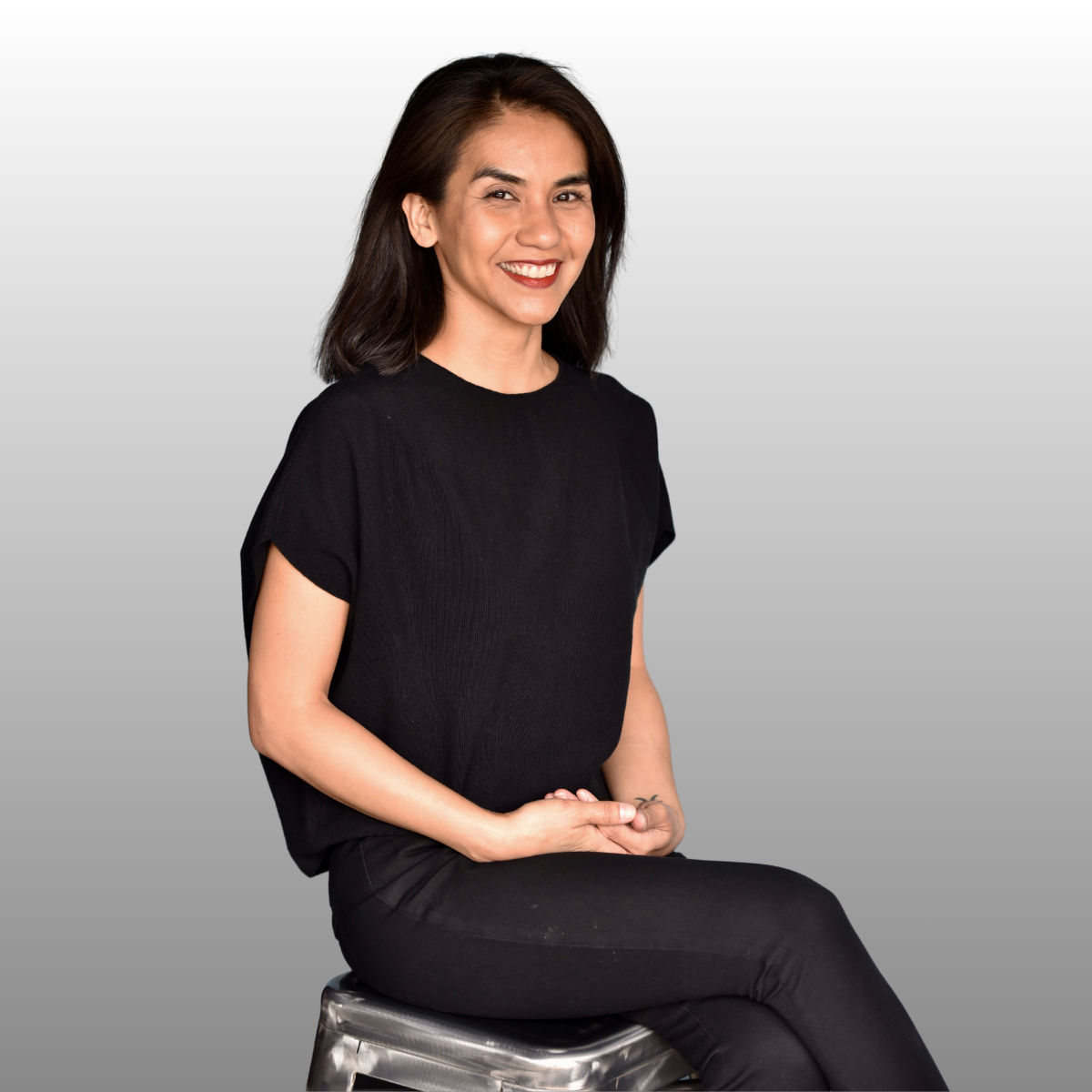 A woman sitting on a silver stool, smiling, wearing a black t-shirt and black pants, with a light gray background.
