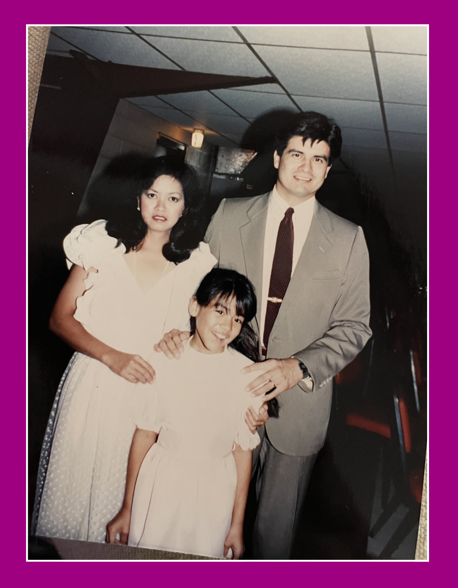 A family of three posing for a photo indoors, with a woman in a white dress, a man in a suit, and a young girl also in a white dress.