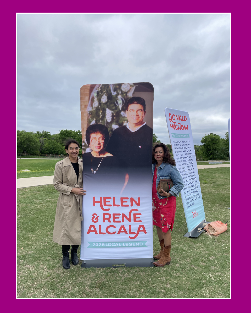 Two women standing outdoors next to a vertical banner that displays a large photo of a woman, with text reading "Helen & Rene Alcala" and "2025 Local Legend". Behind them, there is another banner with information about Donald Muchow. The sky is cloudy, and there are green trees and grass in the background.