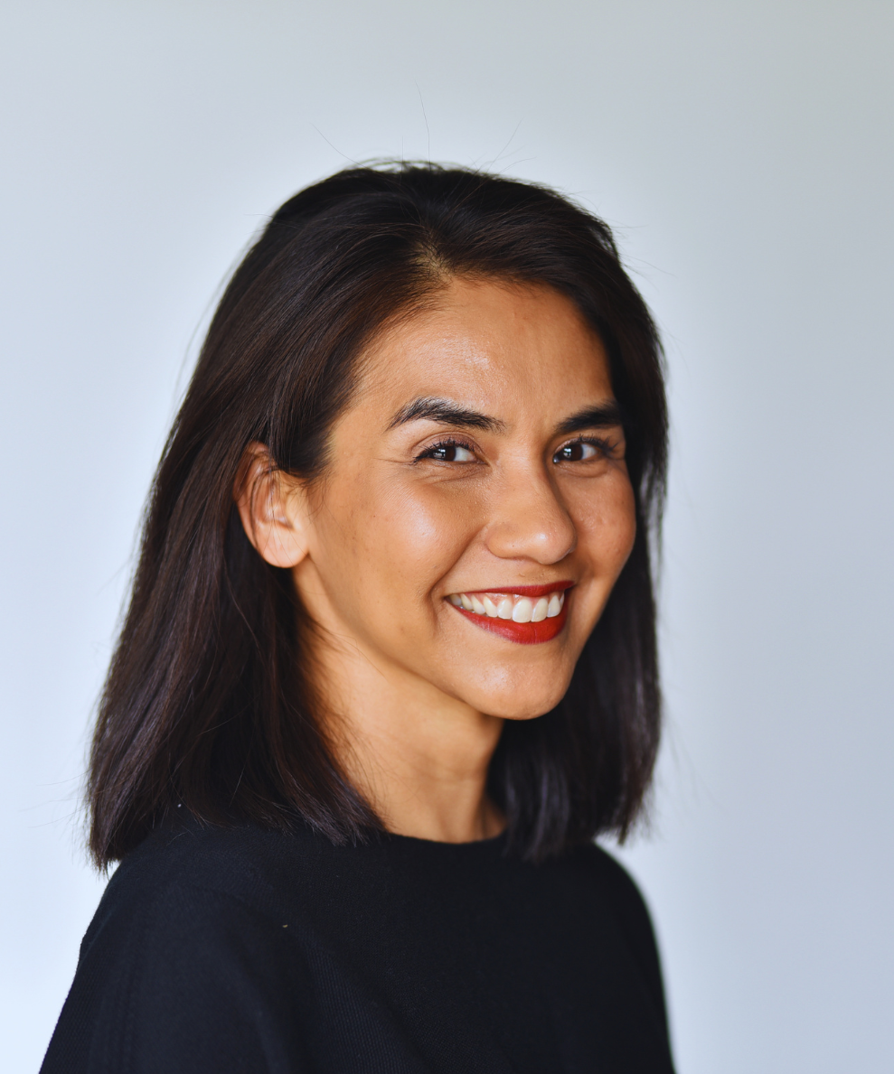 Close-up portrait of a woman smiling, with dark shoulder-length hair, wearing a black top, against a plain light background.
