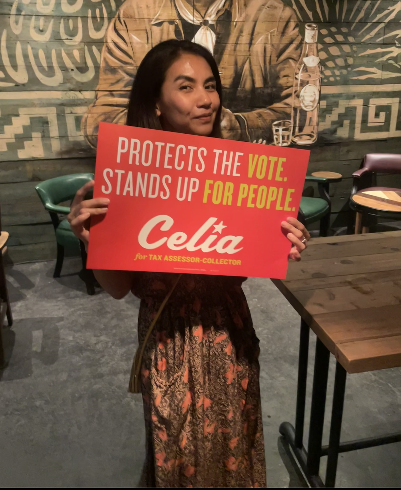 A woman holding a red sign that reads 'Protects the Vote. Stands up for People. Celia for Tax Assessor-Collector' while standing inside a restaurant or cafe. The background features a mural with a person holding a bottle and a glass on a wooden wall.