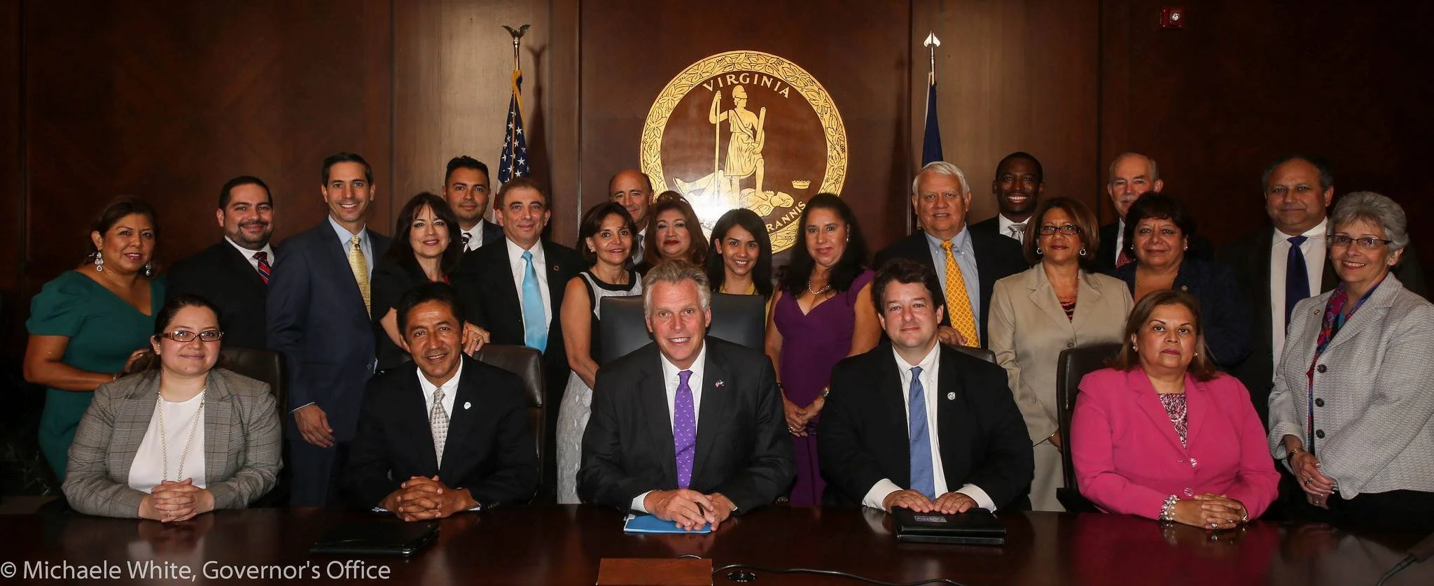Group of people in formal attire seated and standing in a wood-paneled room with a Virginia state seal on the wall behind them.