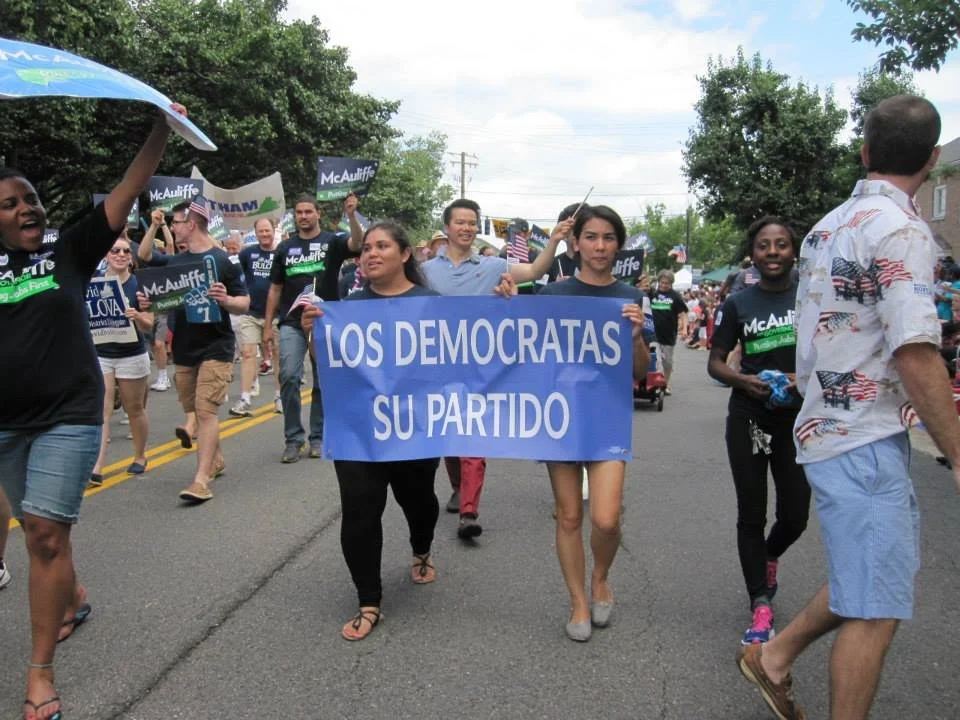 People participating in a parade or march, with some holding a blue sign that reads "LOS DEMOCRATAS SU PARTIDO."