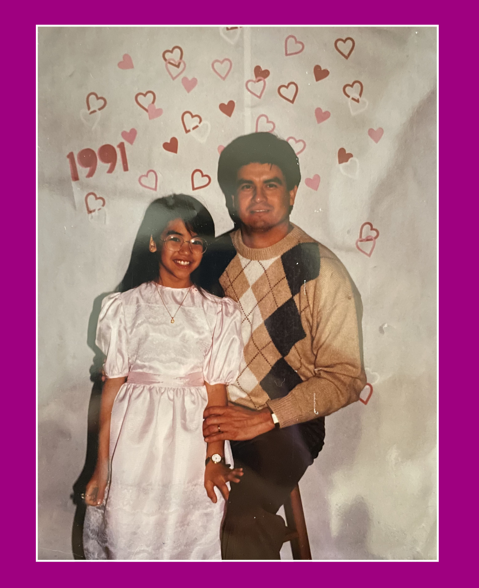 A woman and a man posing in front of a backdrop decorated with pink and red hearts, with the year '1991' written on it. The woman is wearing glasses and a light pink dress, while the man is dressed in a beige sweater with an argyle pattern. They are smiling and holding hands.