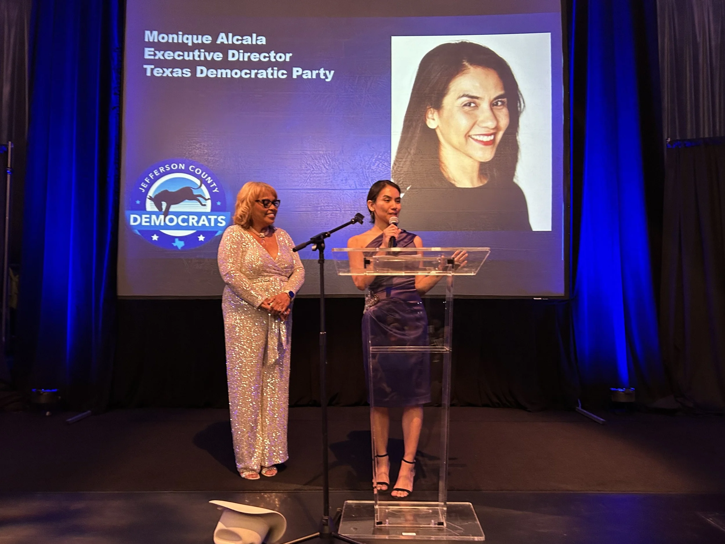 Two women stand on a stage at an event with a large presentation screen behind them. The screen displays a photo of a smiling woman and text identifying her as Monique Alcala, Executive Director of the Texas Democratic Party. There is also a logo for Jefferson County Democrats on the screen.