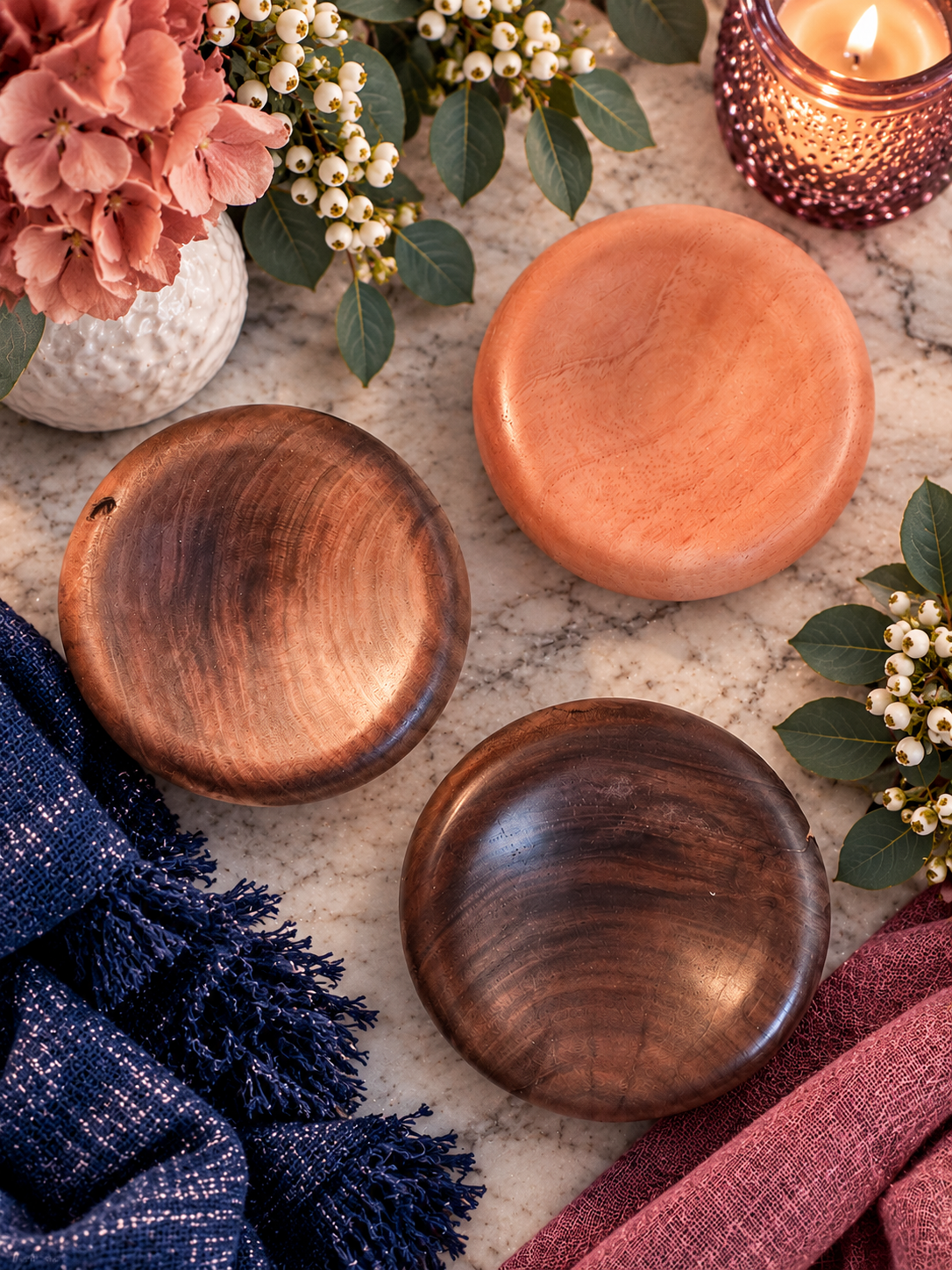 Warm tabletop arrangement with three wooden bowls, coral decor, flowers, white berries, candle, and folded navy and burgundy textiles