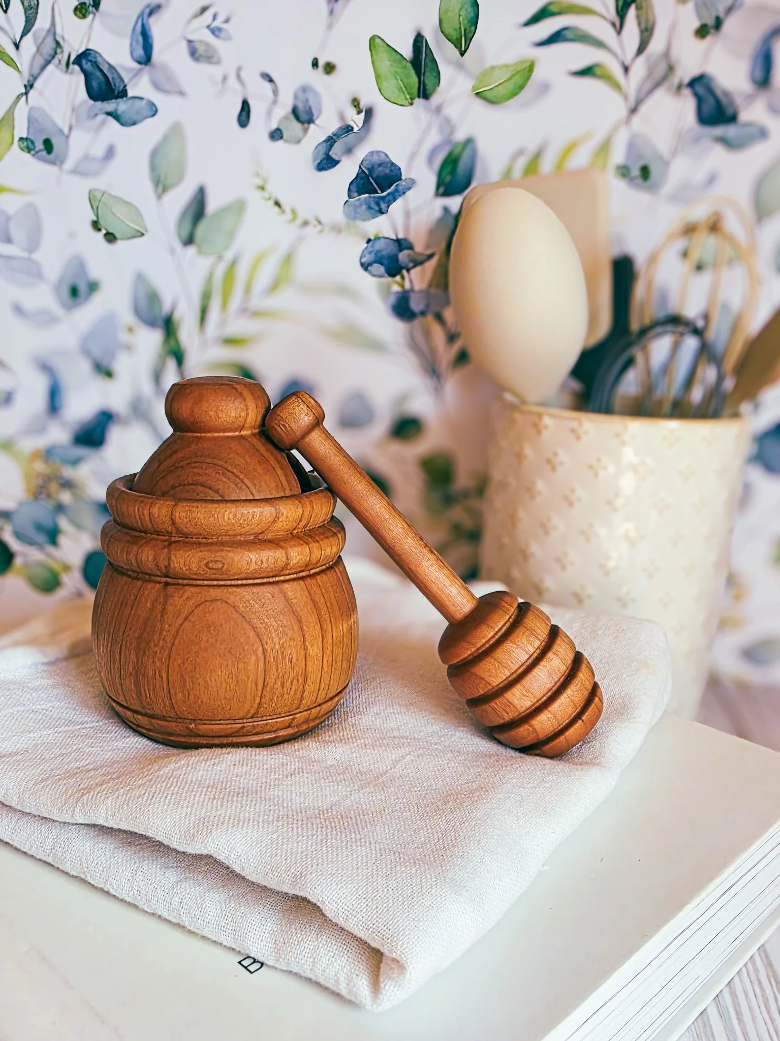 Close-up of a Cherry wood honey dipper resting on a small Cherry wood honey pot, with a background of a white cup filled with utensils and a decorative backdrop of blue and green leaves.