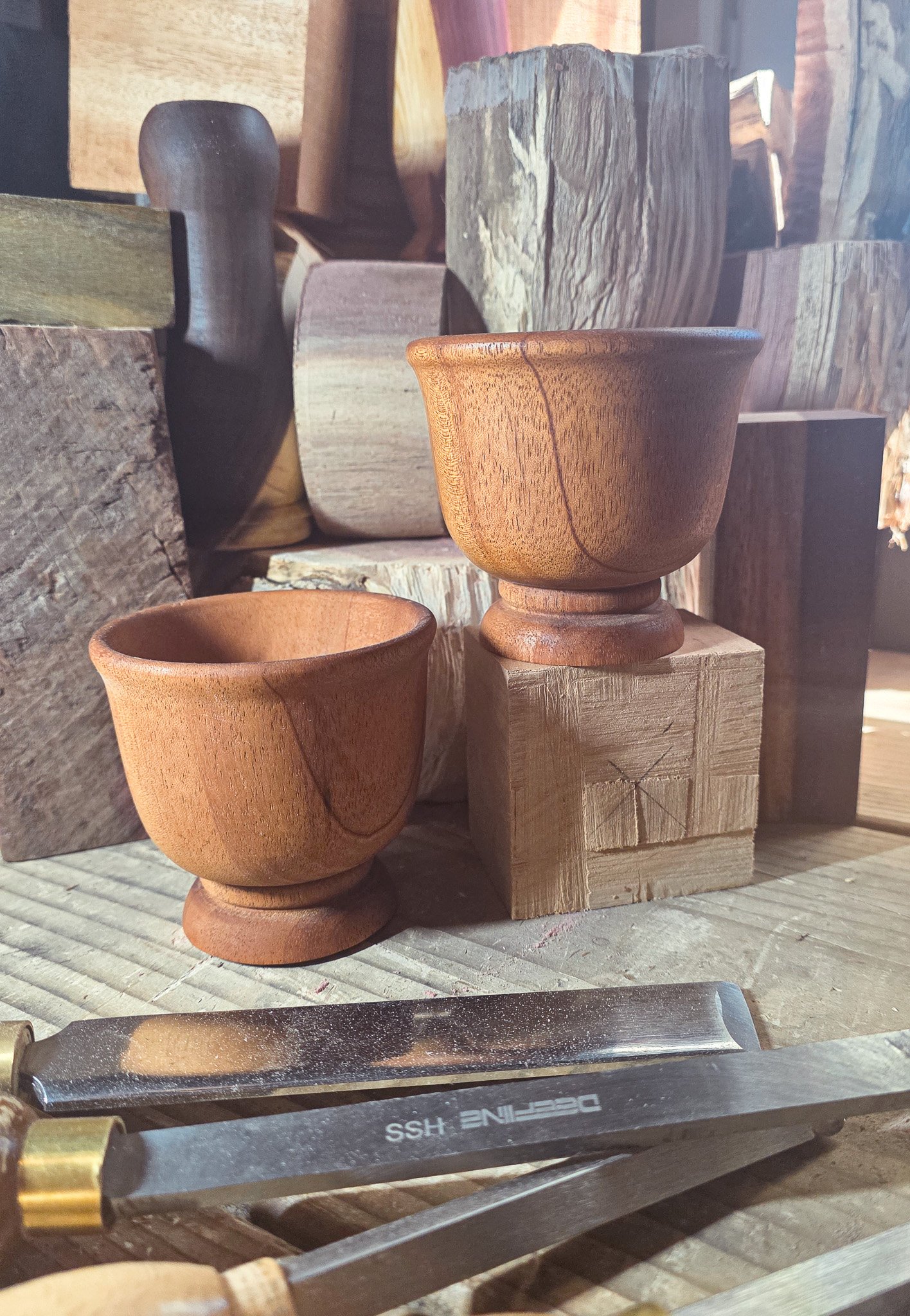 Close-up of wooden bowls turned from African Mahogany and blocks of various wood types arranged on a work surface, with quality woodworking tools nearby, in a woodworking shop or studio.