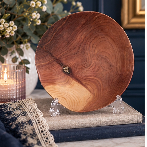 Round live-edge wood slice displayed on a clear stand atop stacked books, with a candle and flowers in the background.