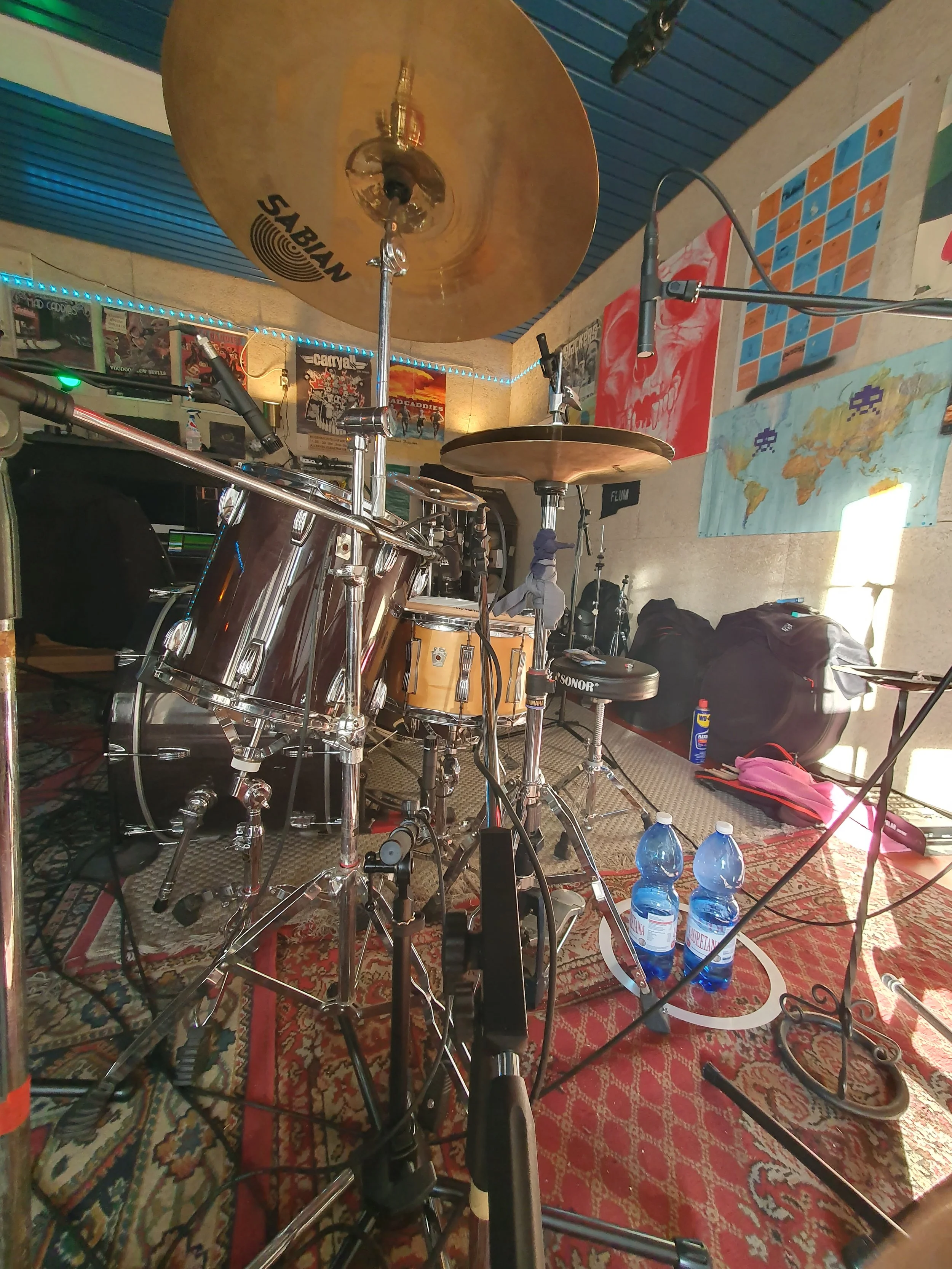Interno di uno studio musicale con batteria, microfoni e palette. Decorazioni alle pareti tra cui poster e una mappa del mondo, con un tappeto sul pavimento.