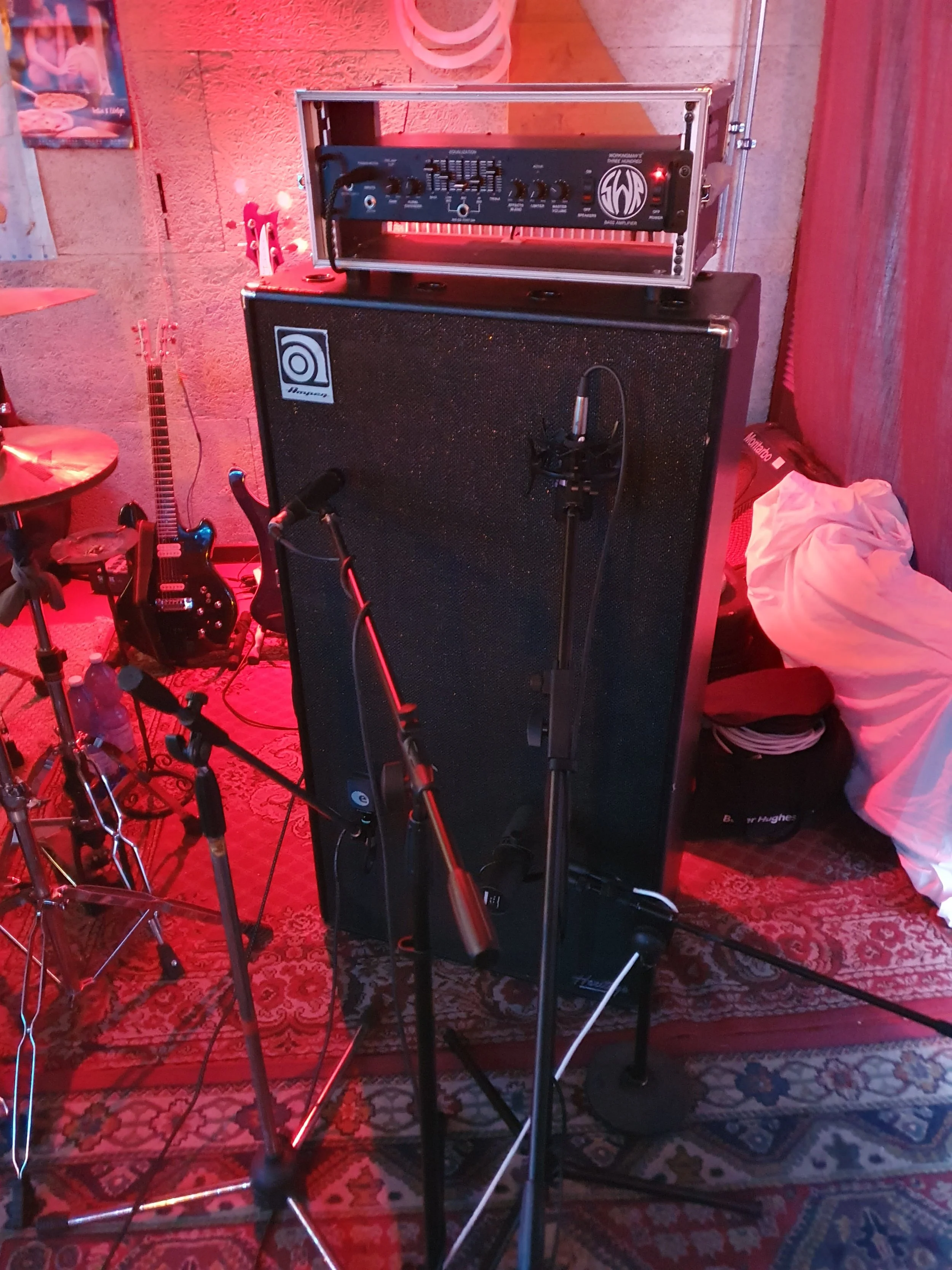 Immagine di attrezzature da musica tra cui una chitarra elettrica, diffusori, un mixer, un microfono e vari cavi in un ambiente con tappeto persiano e luci rosse.