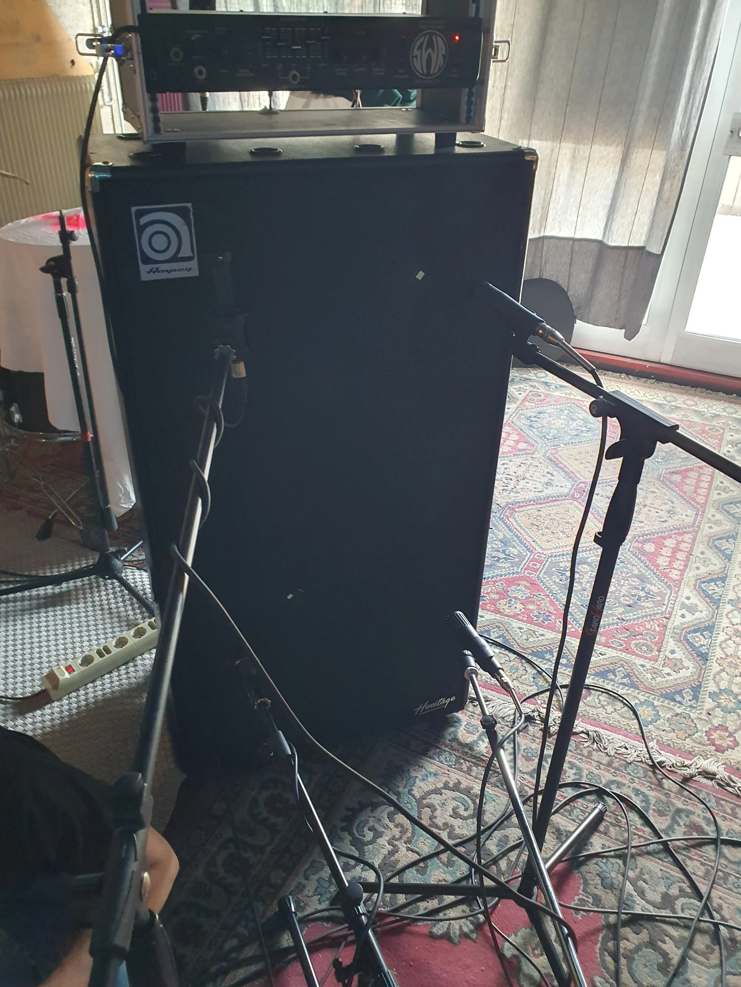 Settore di attrezzature audio con amplificatori, microfono, cavi e supporti, in una stanza con tappeti e tapparelle.