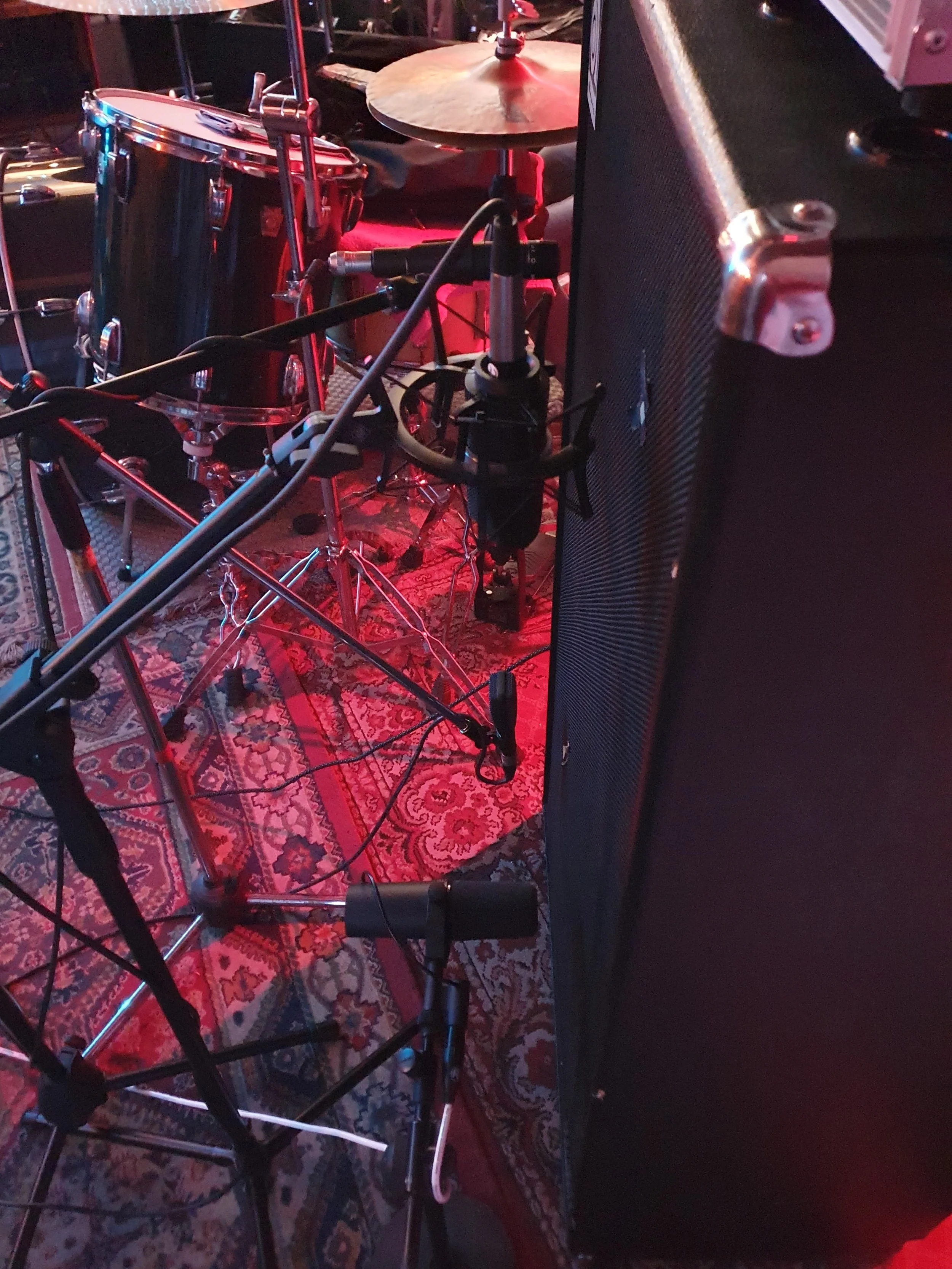 Immagine di una batteria, con vari piatti e timpani, in un ambiente di musica dal vivo, con un grande amplificatore nero in primo piano sulla destra e un tappeto orientale con motivi floreali.