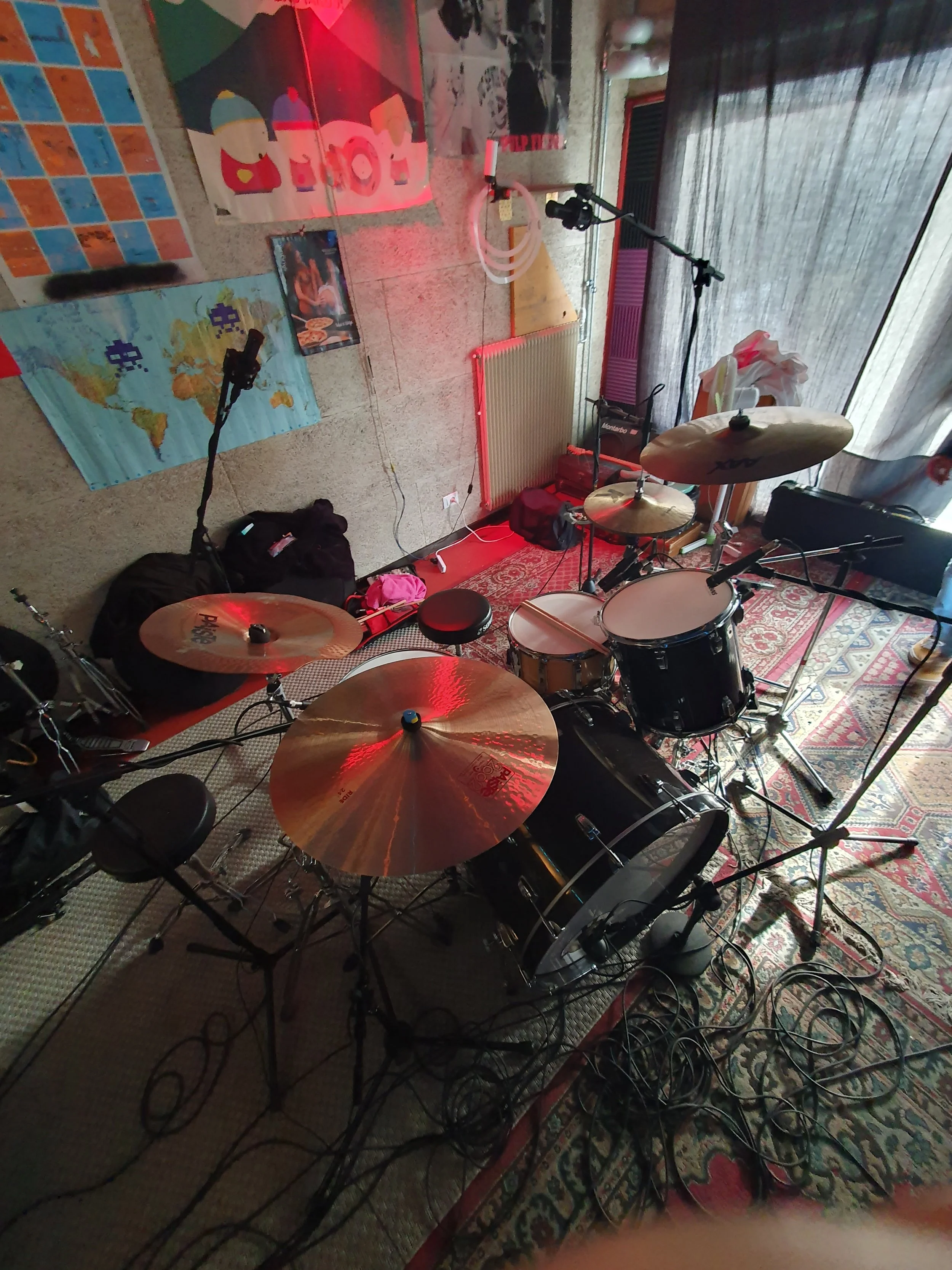 Una stanza con strumenti musicali come batteria e microfoni, decorata con poster e tappeti, con apparecchiature audio e cavi disposti sul pavimento.