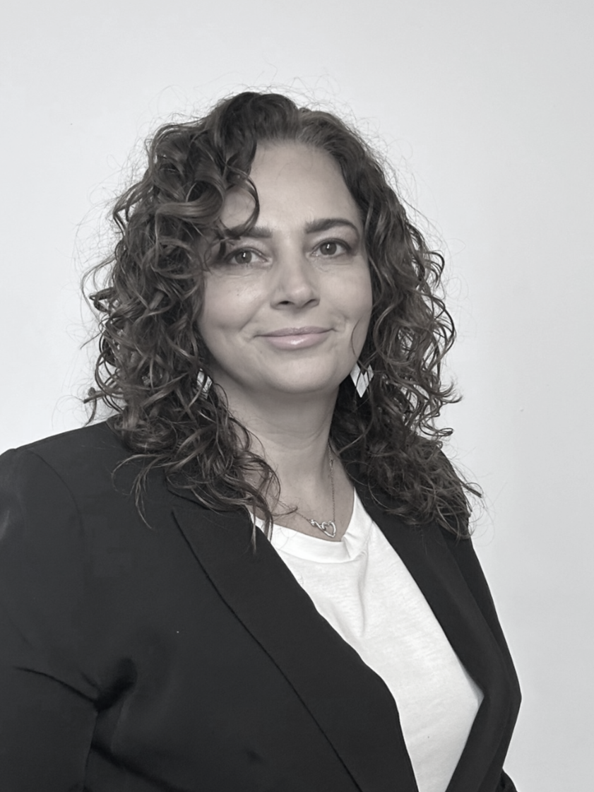 A female hairstylist  with curly hair wearing a black blazer, white shirt, and jewelry, standing against a plain light background.