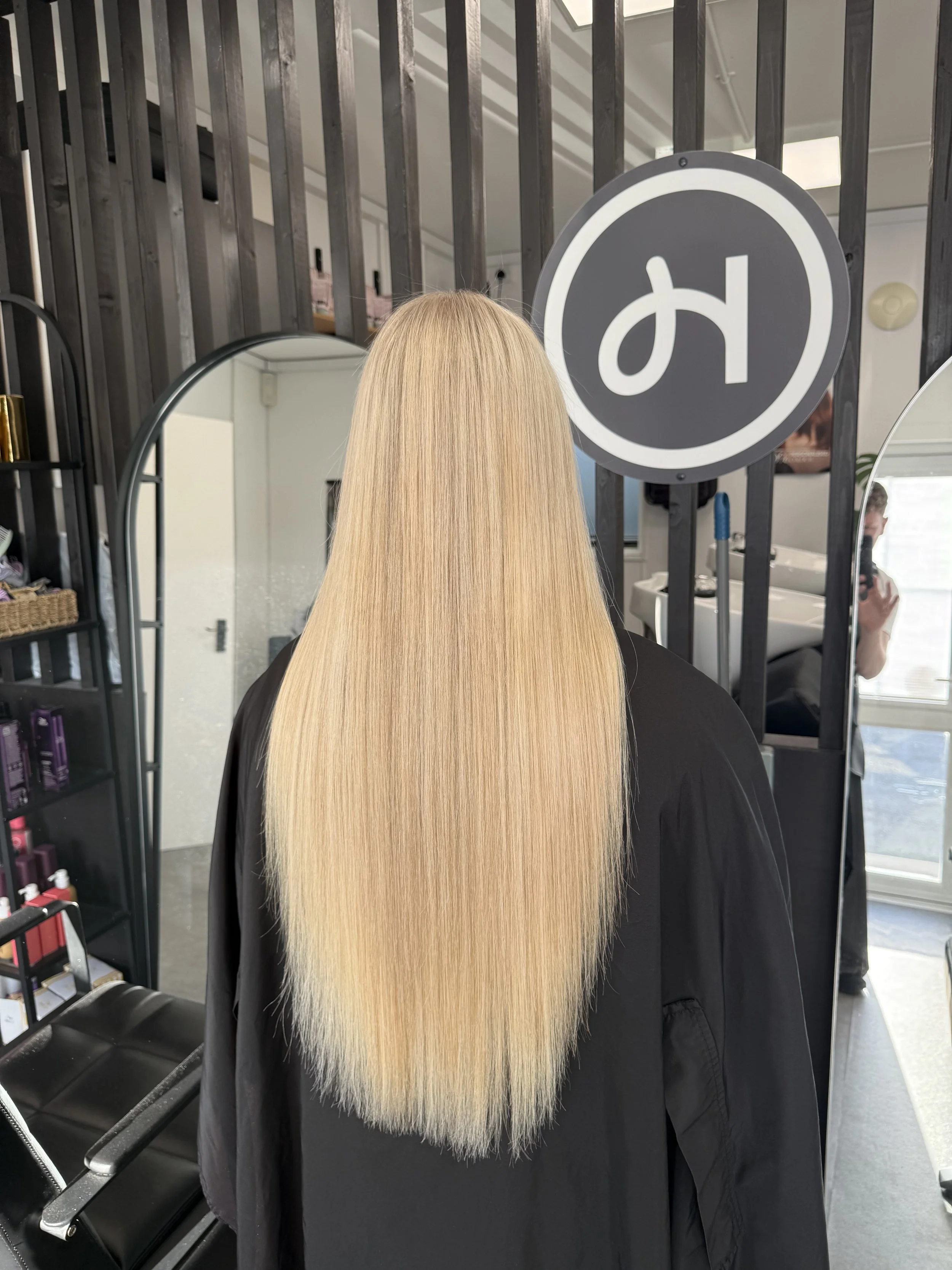 A person with long, straight blonde hair standing in front of a mirror in a salon, facing away. There is a black sign with a white lowercase letter 'h' on it hanging on the wall.