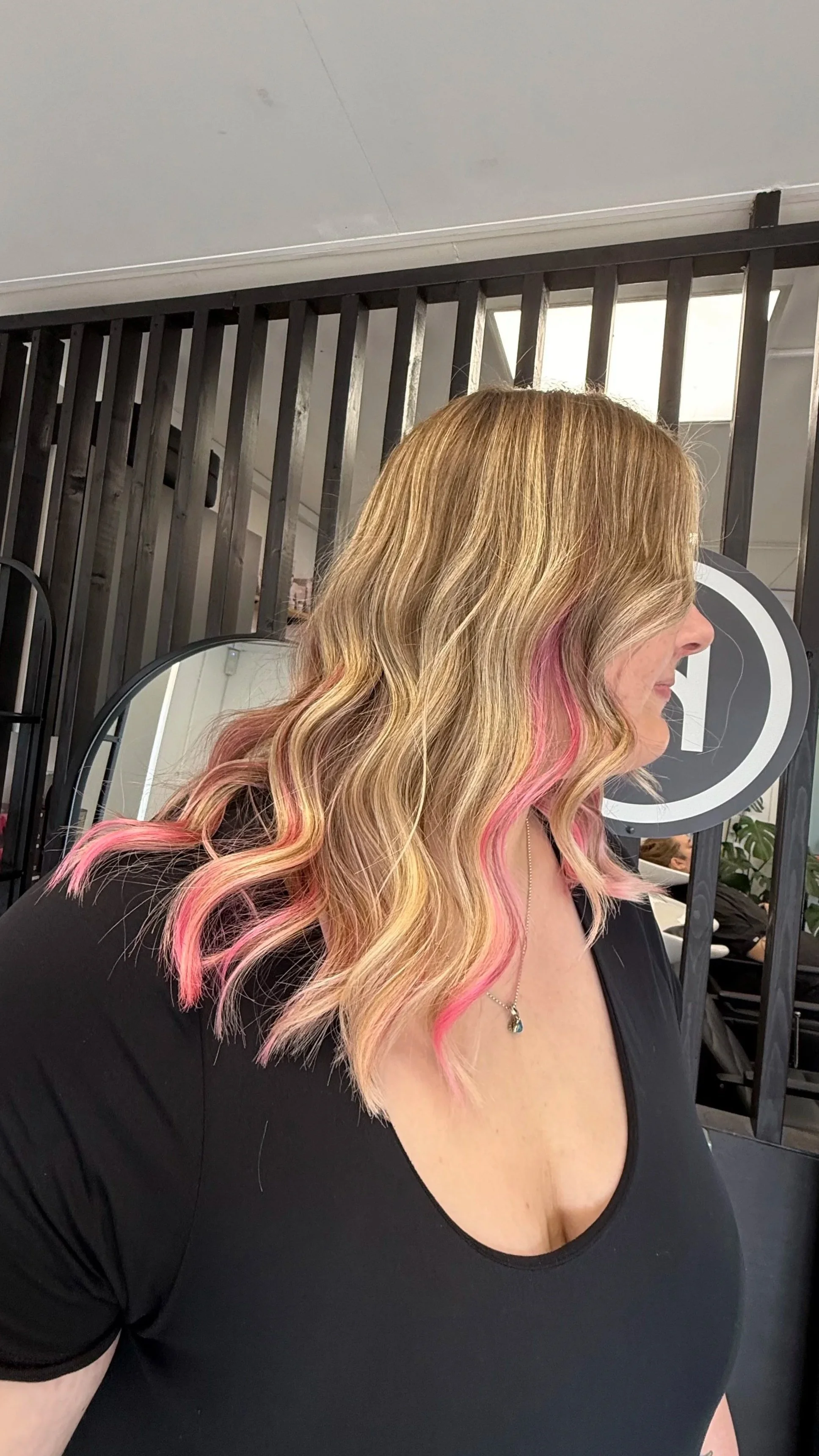 A woman with blonde hair styled in waves, with pink highlights, wearing a black top and a pendant necklace, sitting in a hair salon.