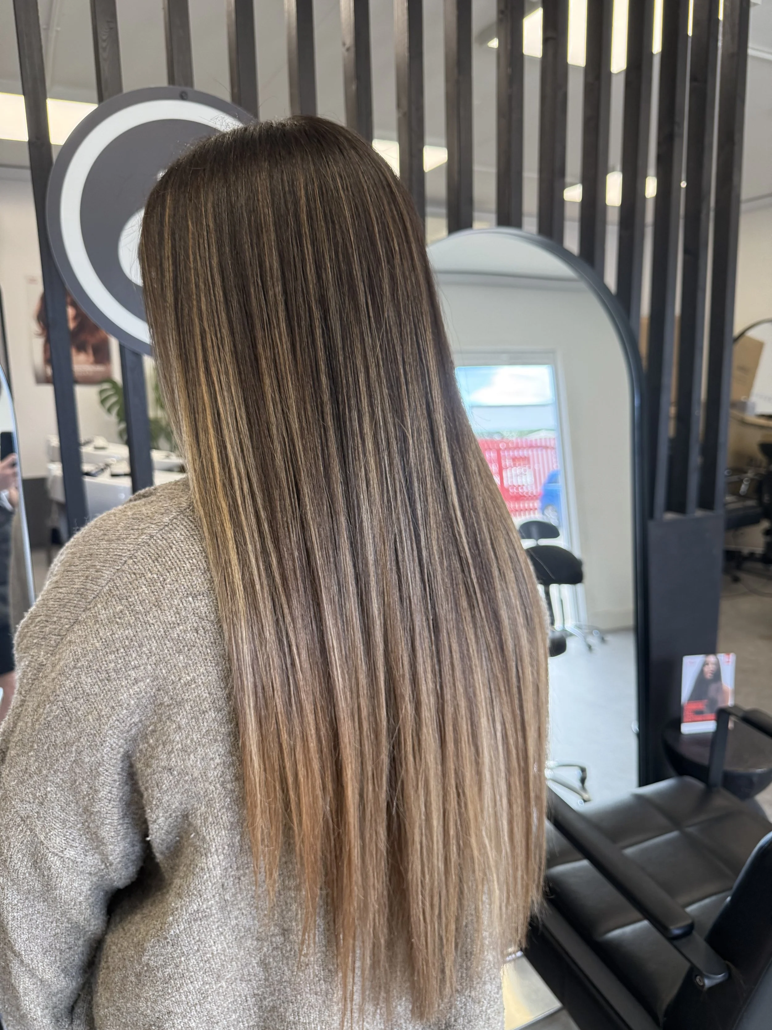 Person with long, shiny, straight hair in a salon or beauty shop, standing in front of a mirror, with salon chairs visible in the background.