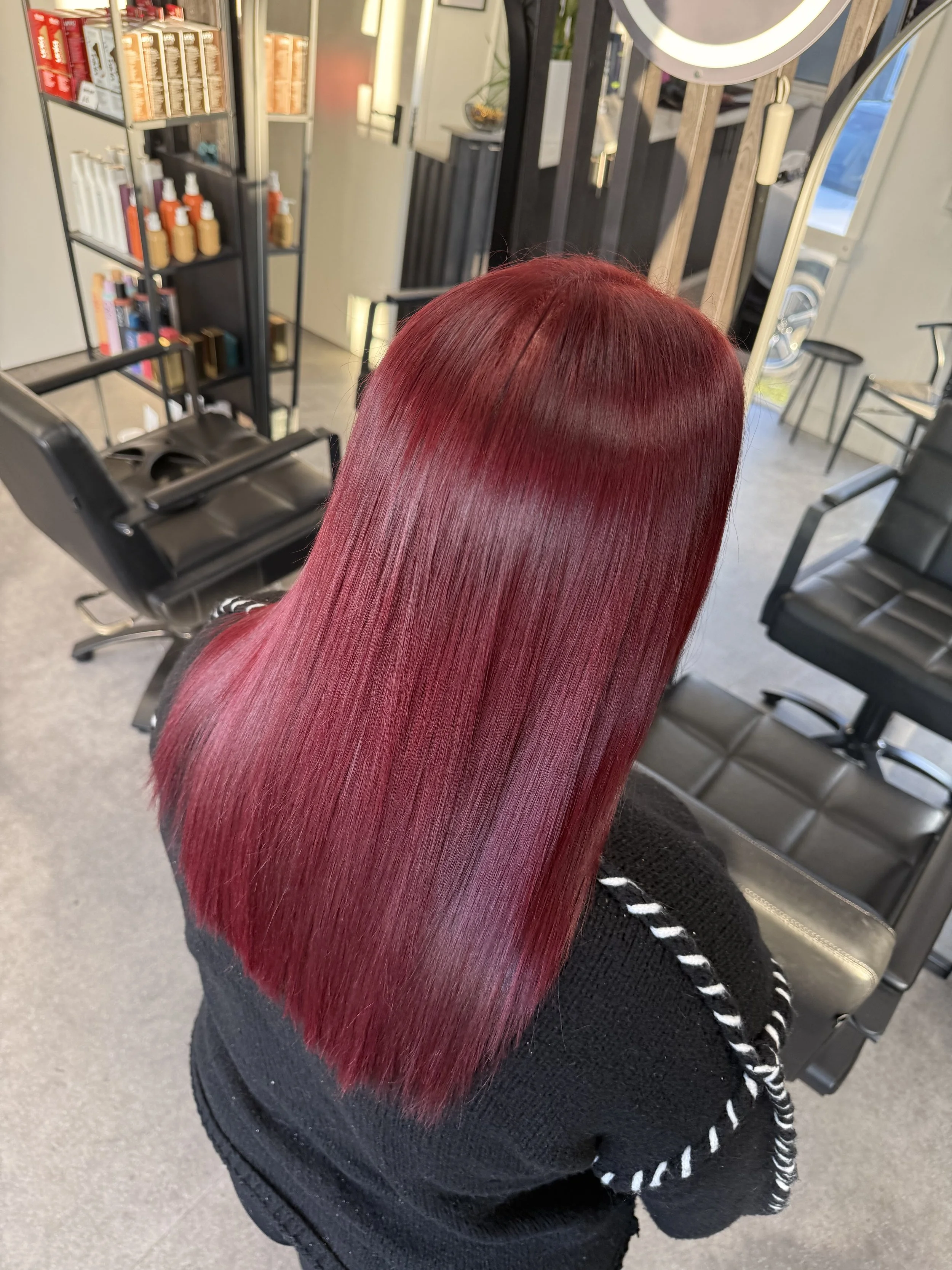 Person with long, straight, vibrant red hair sitting in a salon chair. The salon has a mirror, shelves with hair products, and other salon chairs.