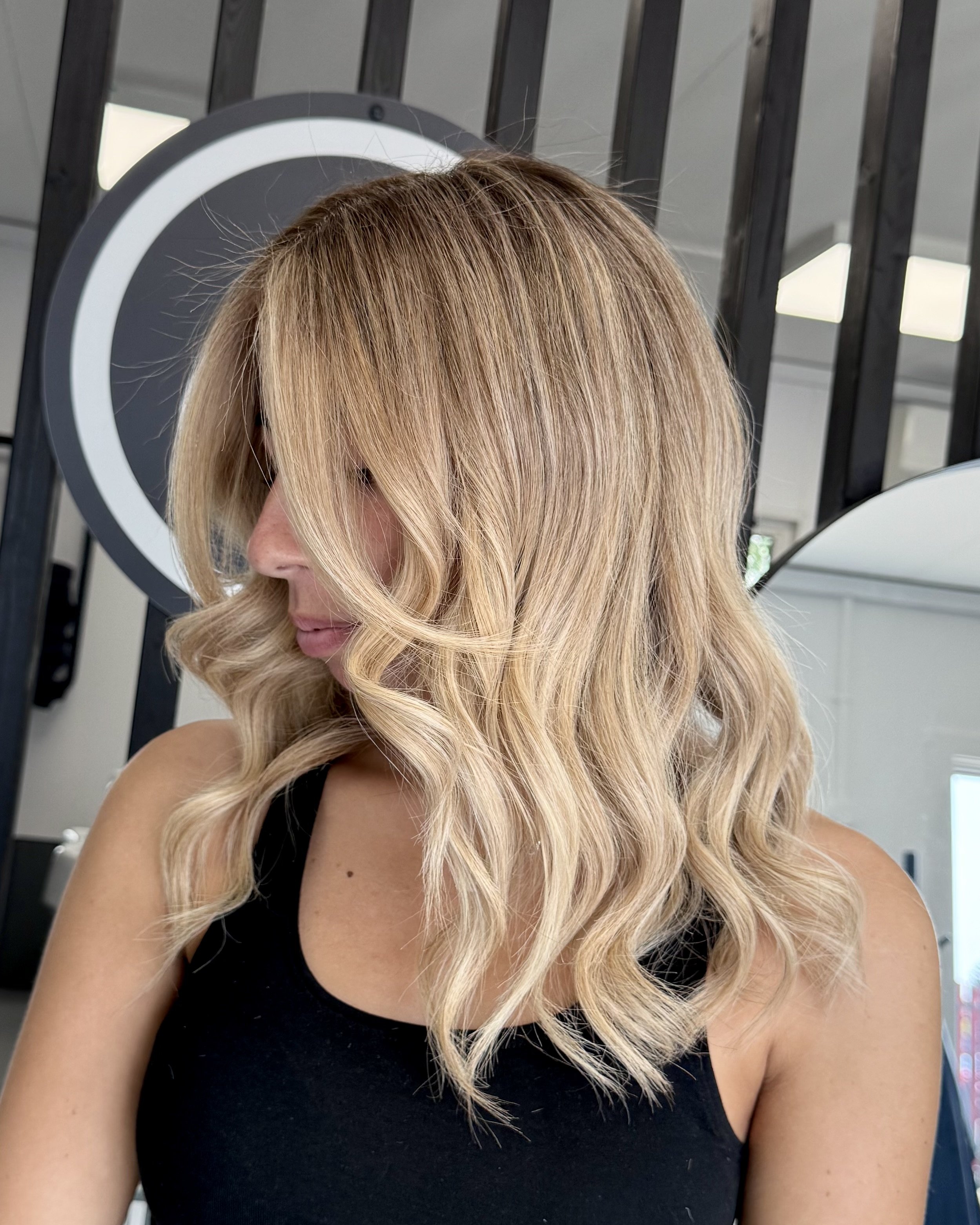 Close-up of a woman with blonde, wavy hair and wearing a black sleeveless top in a salon or studio setting.