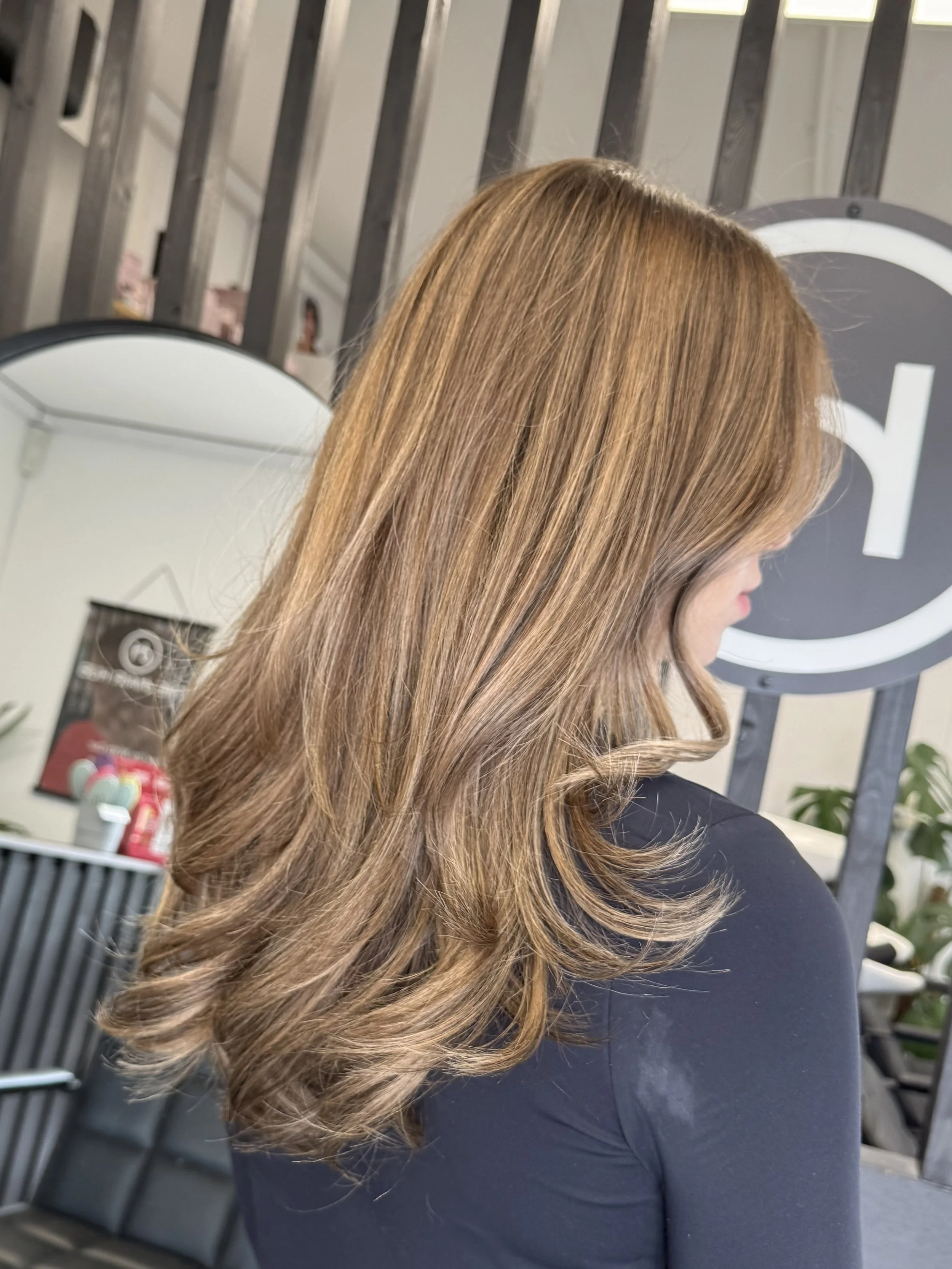 A woman with long, wavy, light brown hair wearing a black top, standing indoors in front of some mirrors and a gray slatted wall, with a salon or beauty studio setting.