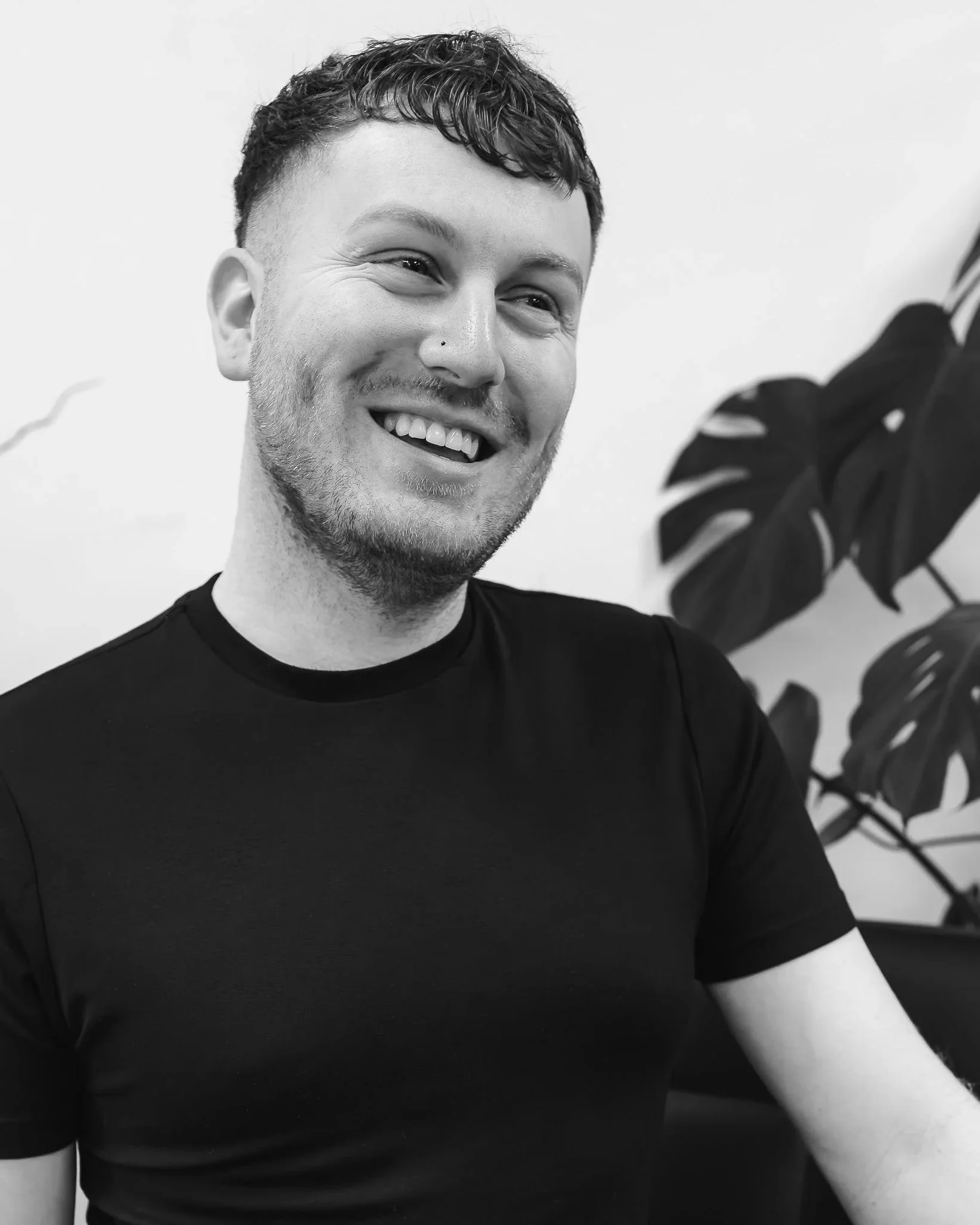 A male hairstylist with short, curly hair and a nose piercing, wearing a black t-shirt, standing in front of a plain wall with a plant partially visible on the right side.