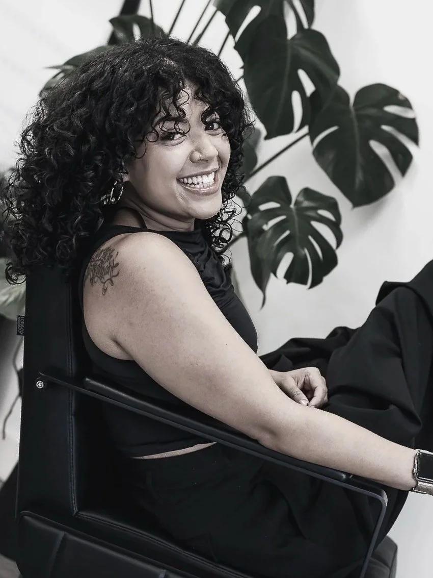 A female hairstylist with curly dark hair smiling, sitting in a black chair, with large monstera leaves in the background.