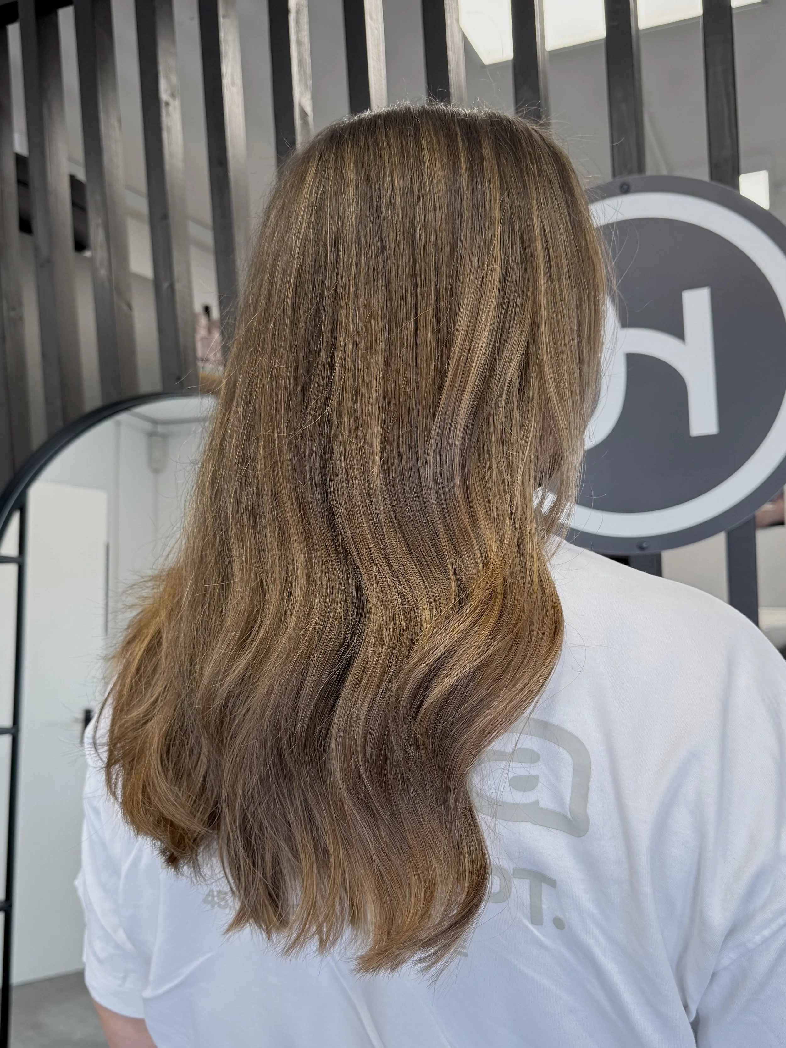 A person with long, brown hair wearing a white shirt, standing in front of a mirror and a black sign with white letters.