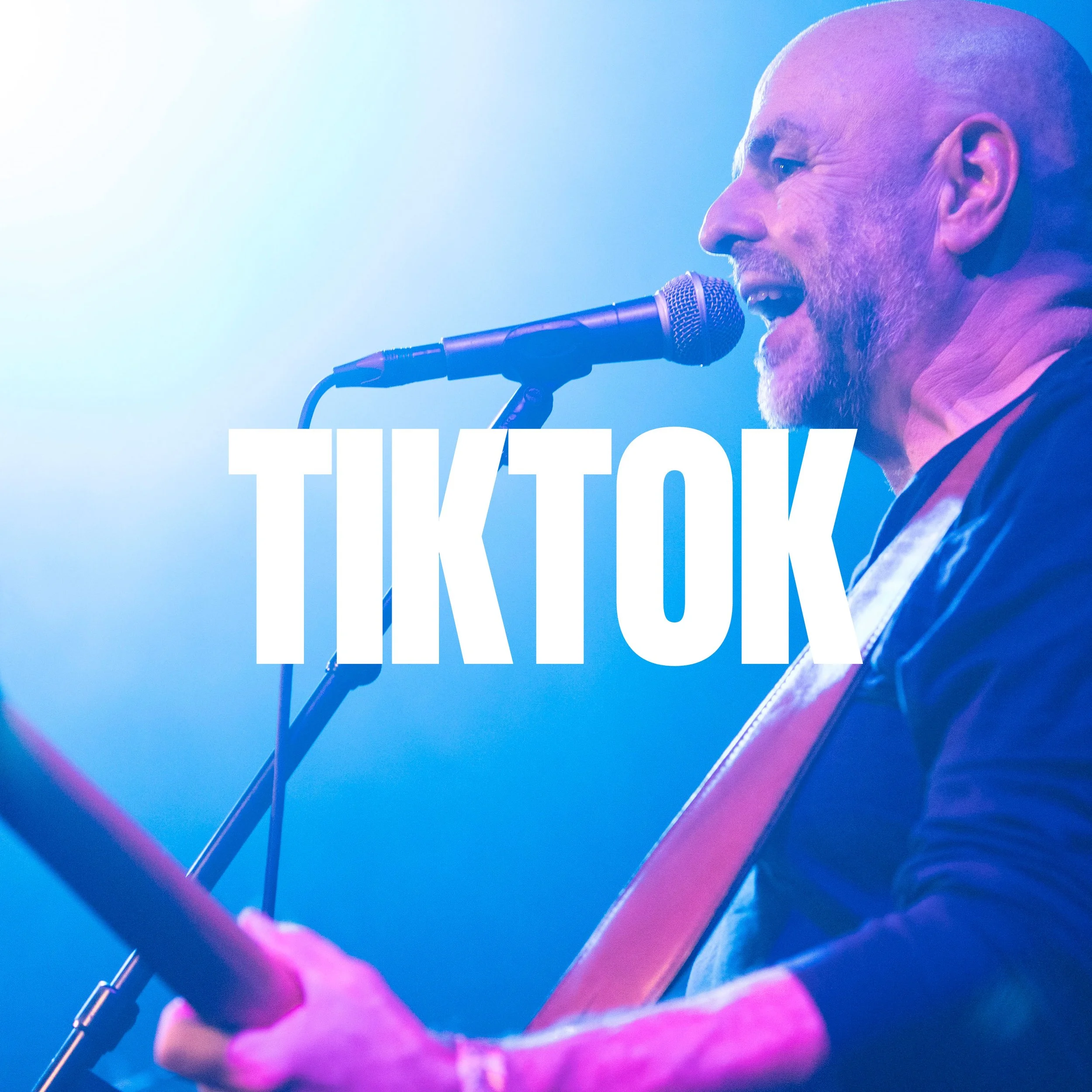 A man with a beard singing into a microphone on stage with blue and purple lighting, partially obscured by pink stage equipment.
