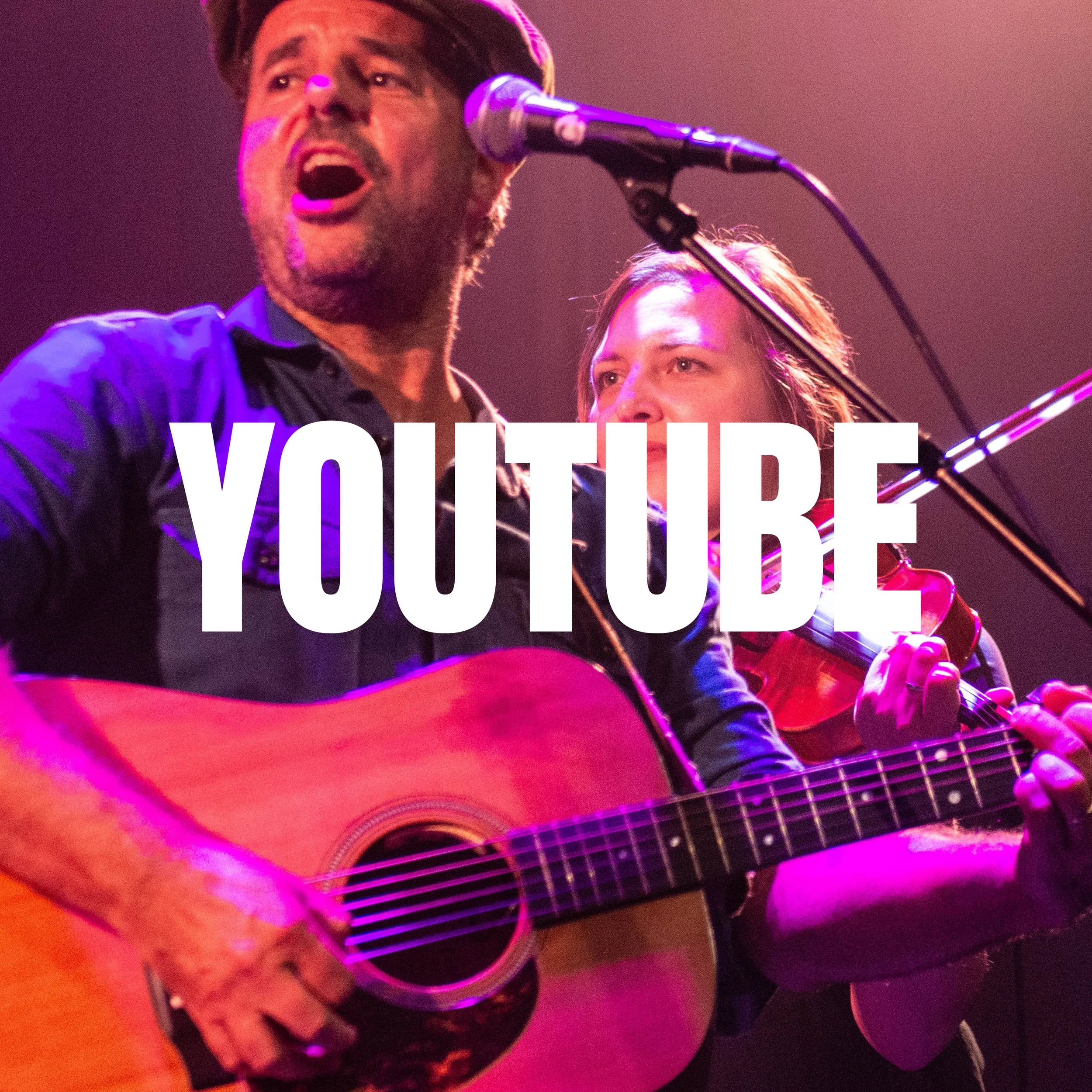 Dieter van der Westen singing into a microphone on stage, playing an acoustic guitar, with a woman beside him also playing guitar, illuminated by purple stage lighting.