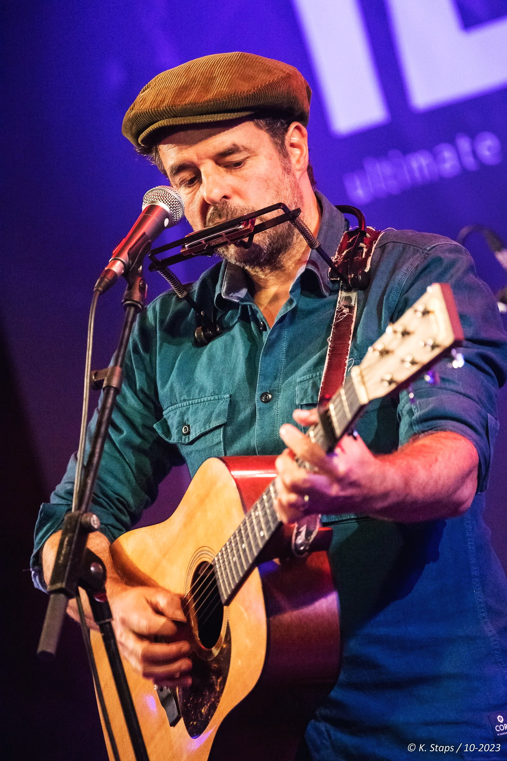 Dieter van der Westen wearing a brown cap playing an acoustic guitar on stage, with a microphone in front of him and a purple background with text.