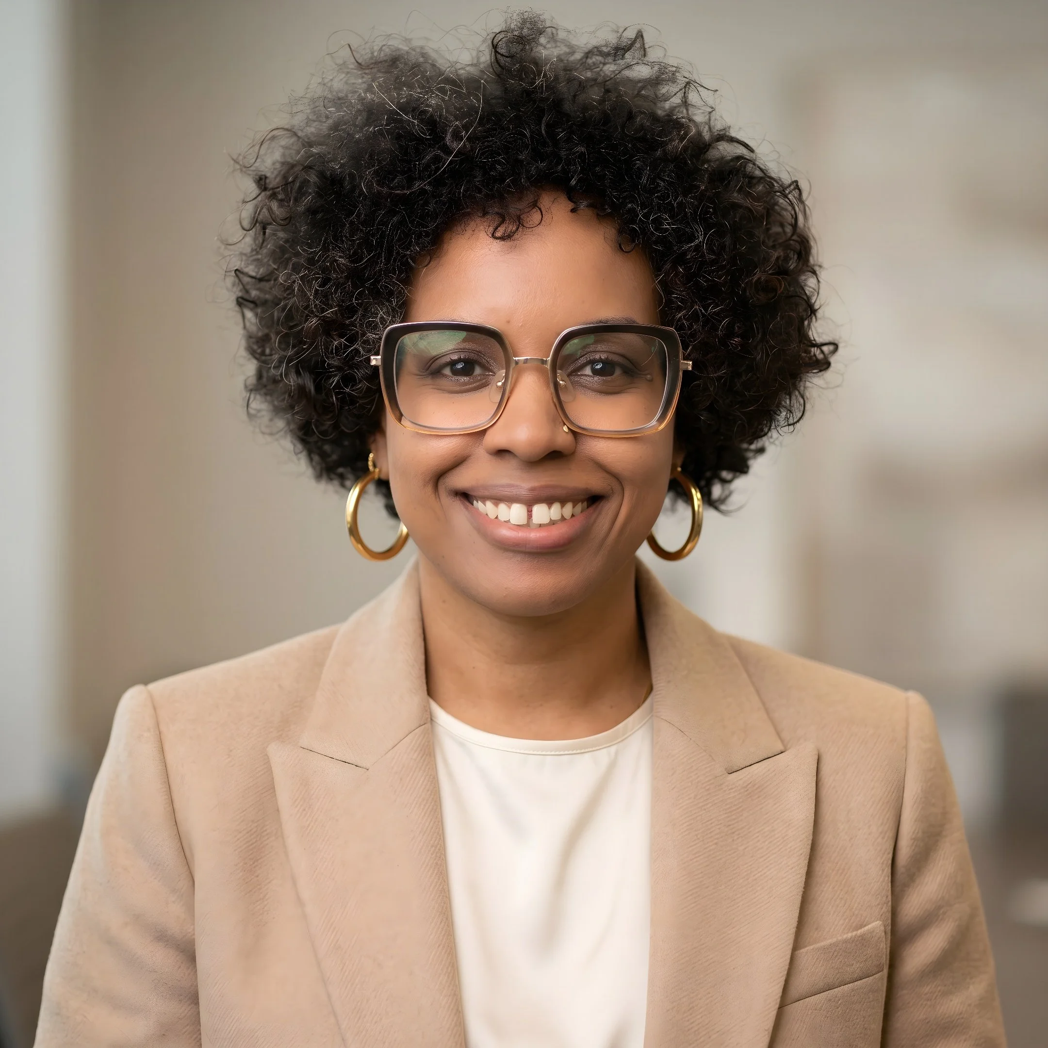 Close-up of a woman's face with curly black hair, wearing large glasses, gold hoop earrings, and a beige blazer over a white top, smiling.