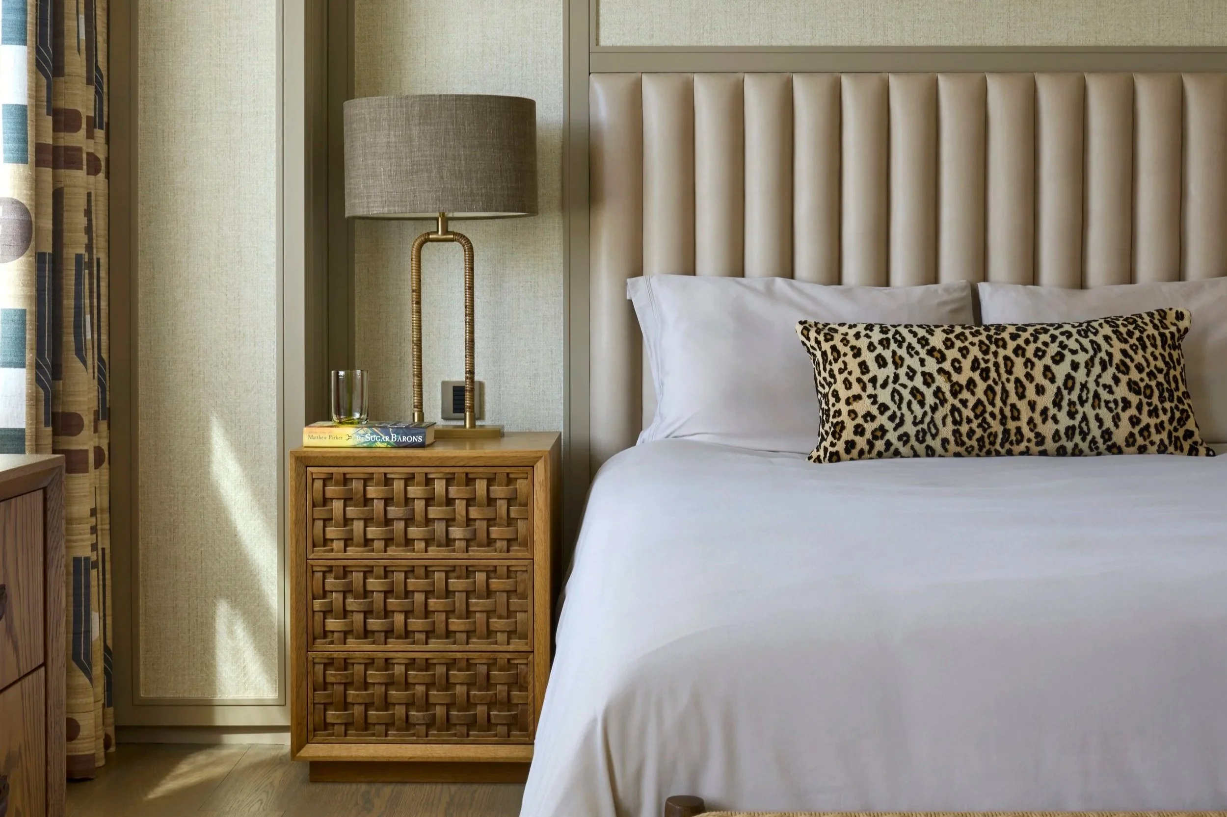 Close-up of a hotel bed with a white pillow and a leopard print pillow, beige headboard, and side table with a lamp, books, and glass of water.