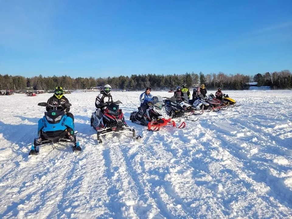 Six people wearing helmets and winter gear sit on snowmobiles in a snowy field with trees in the background.