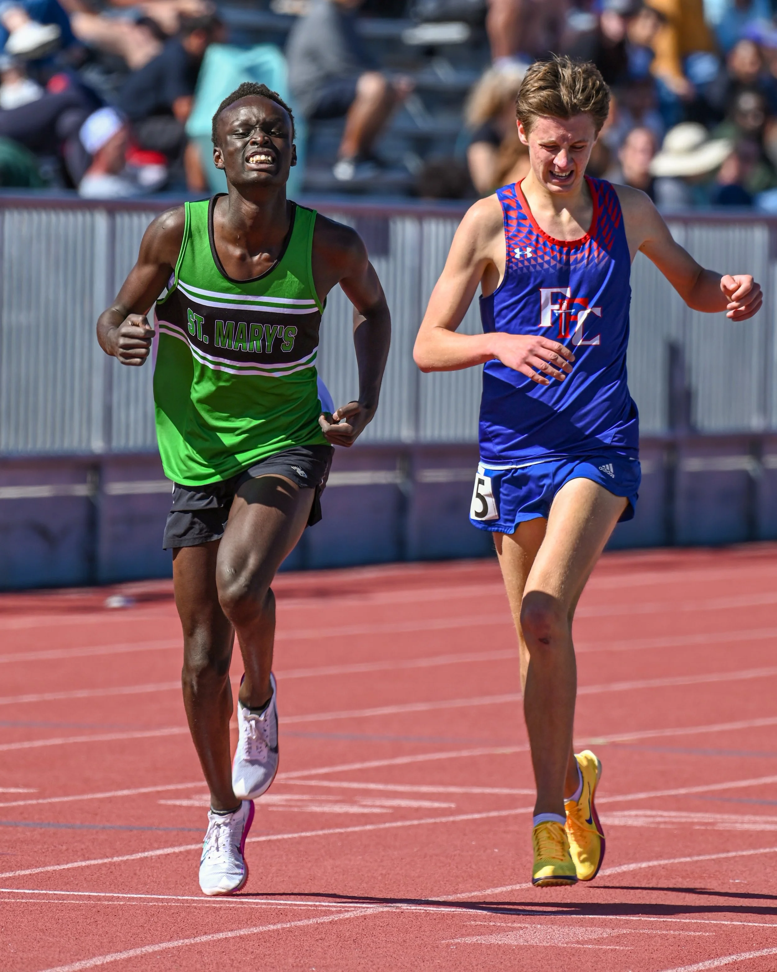 Two male athletes running on a red track during a race, with a blurred crowd of spectators in the background.
