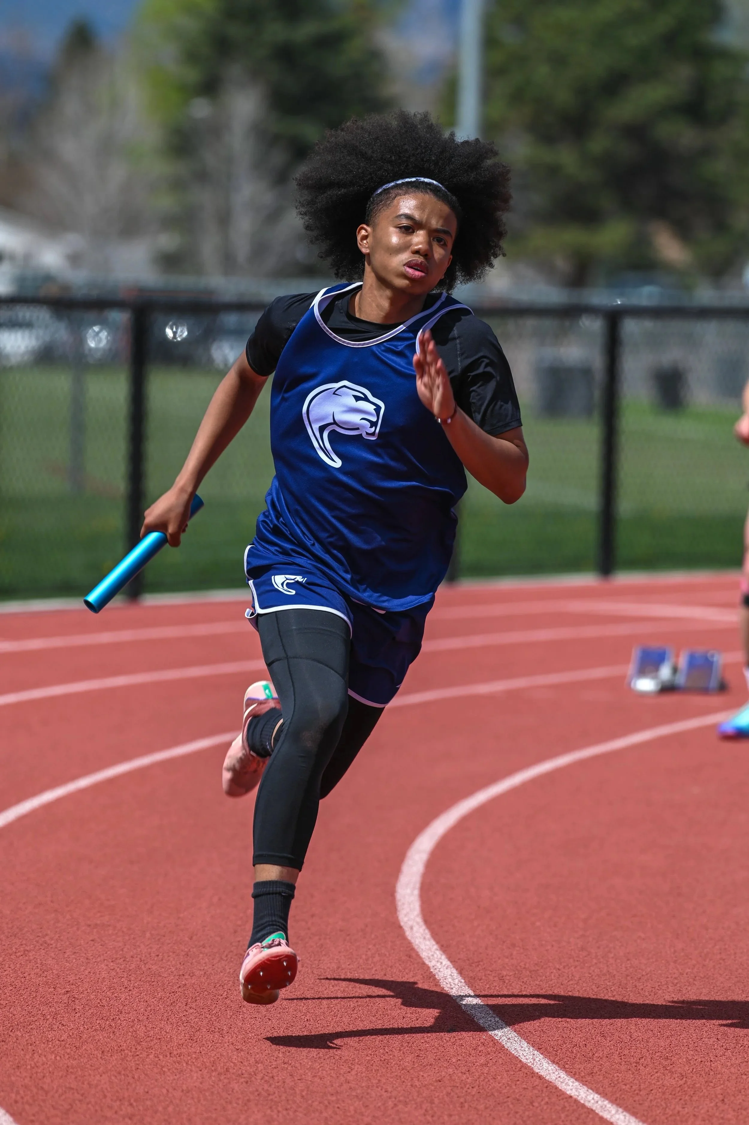 A male high school runner sprints around a curve while carrying a relay baton during the 4x200m relay at a track & field meet.