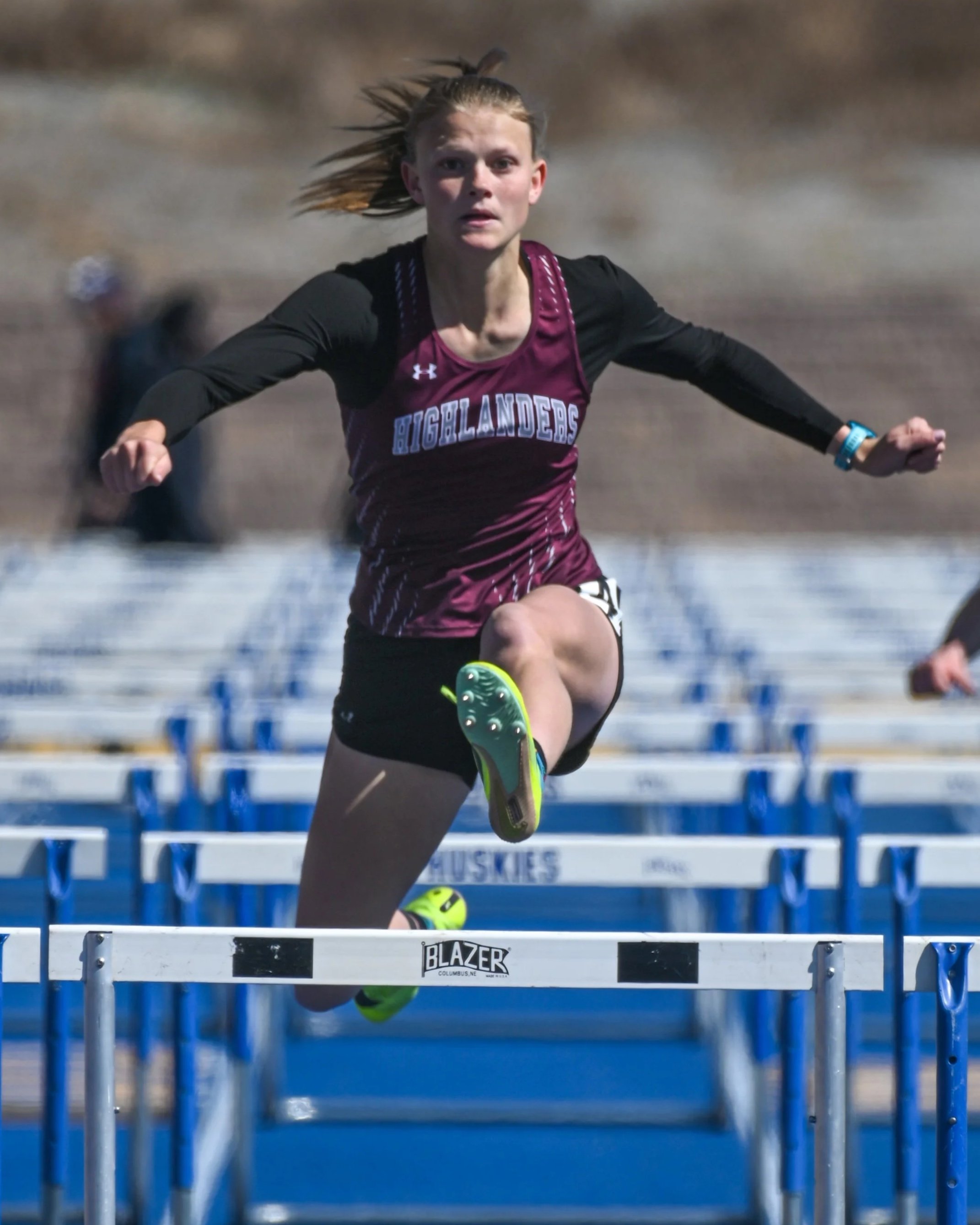 A female high school athlete with long hair running on a blue track, clearing hurdles with a determined expression. 100m hurdle event during a high school track & field meet.