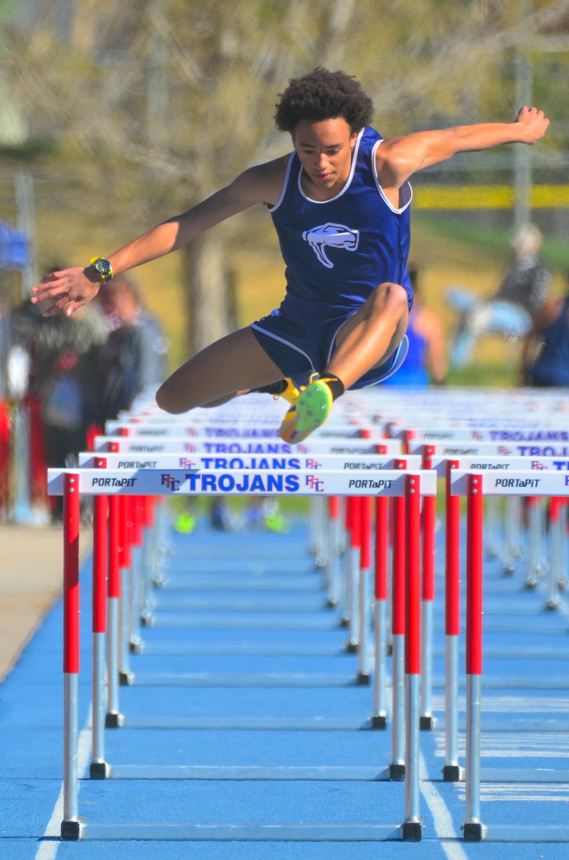 Young male athlete in blue sports uniform and yellow running shoes clearing hurdles in a track race, with spectators blurred in the background.