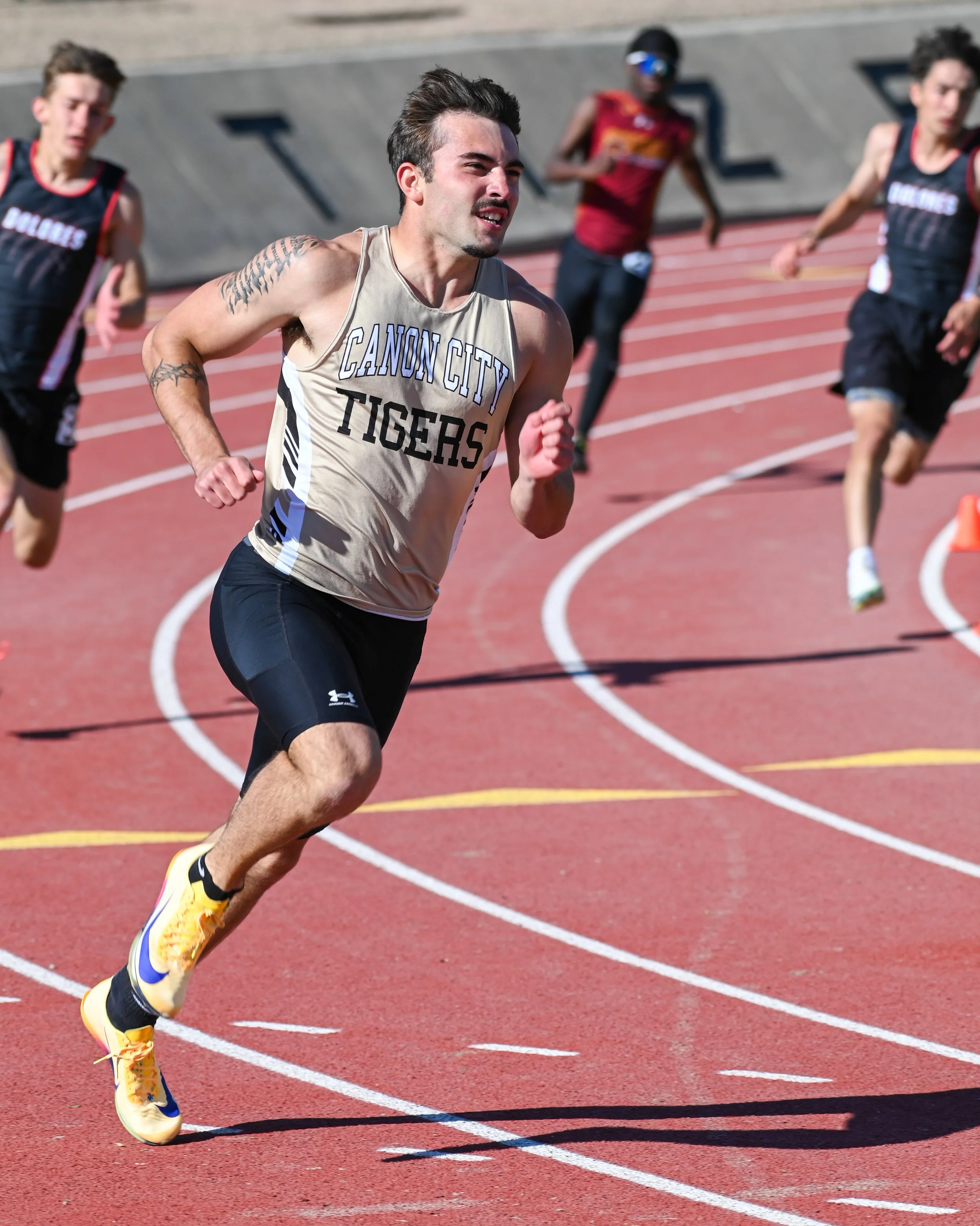Male sprinter wearing a Cañon City Tigers shirt running on a red track with white lane lines during daytime, with other runners in the background.