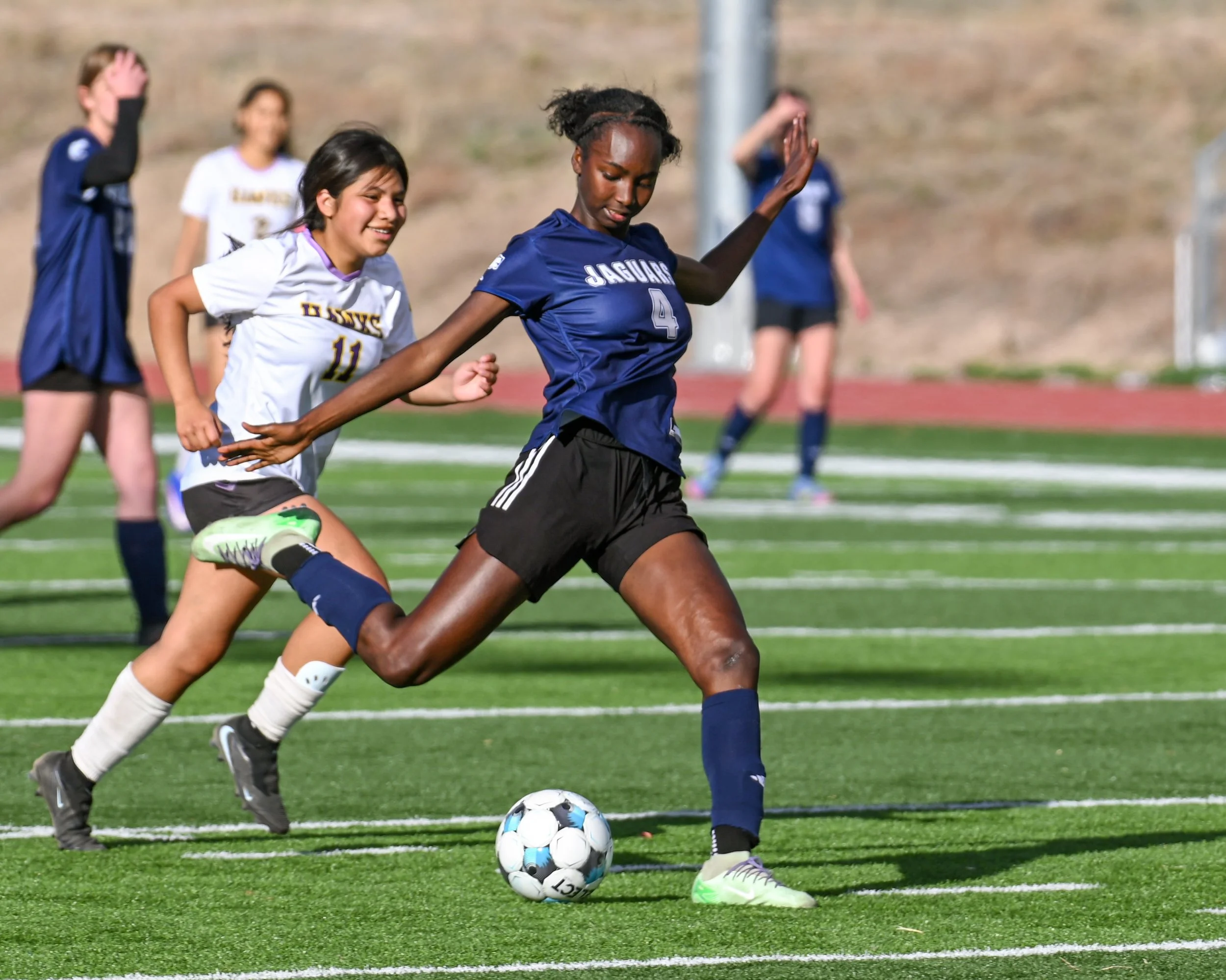 Photograph of a girls' soccer game with players in blue and white uniforms competing for the ball on a green field. The female player in blue is preparing to kick a shot on goal.