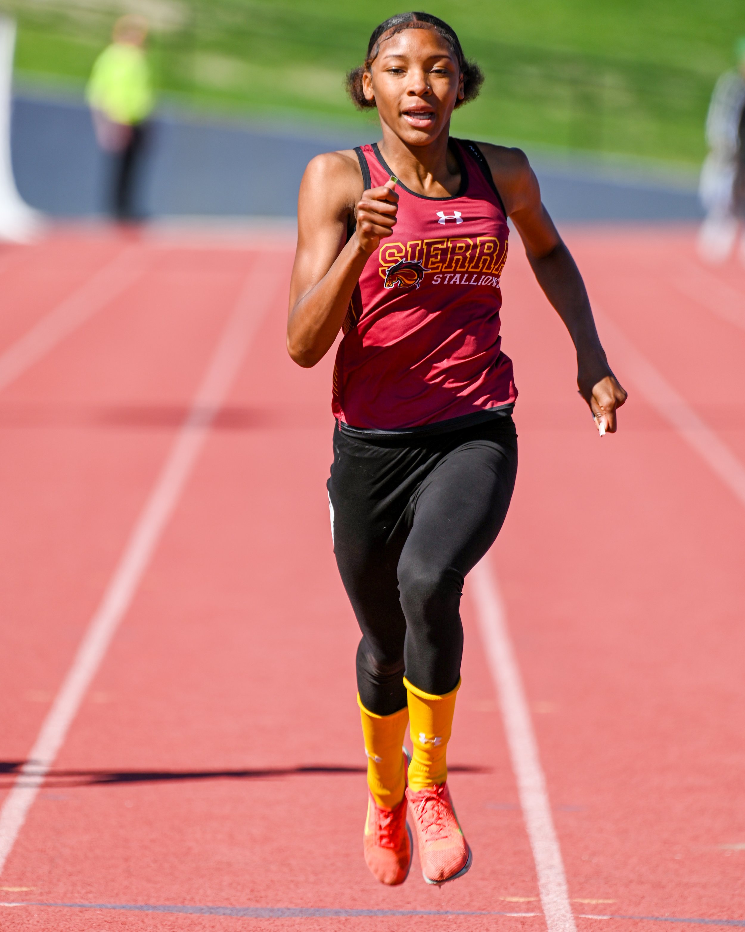 Young female runner in a maroon jersey running on a red track with a green field in the background.