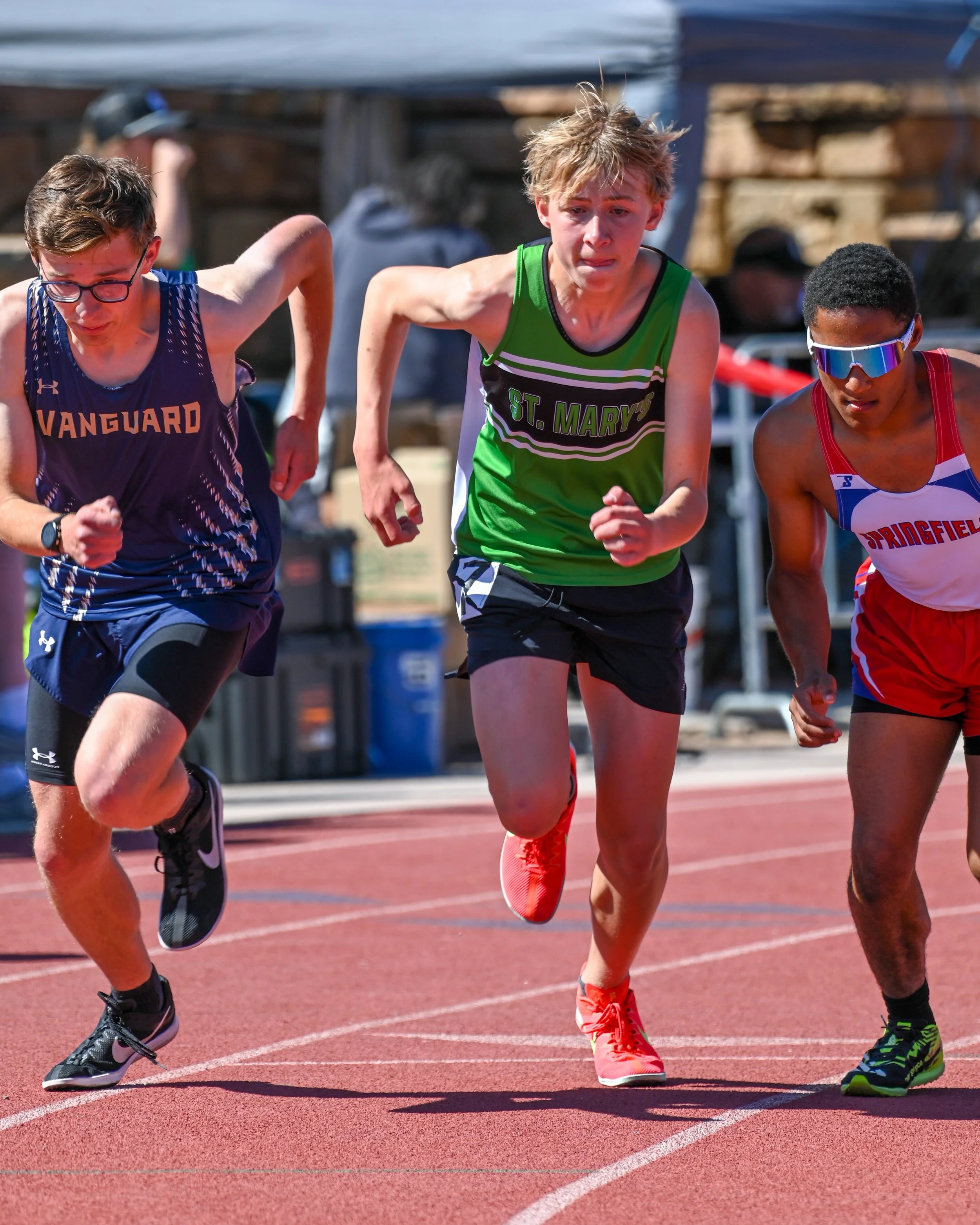 Three male track athletes running on a track at the start of a distance event, focused and in motion.
