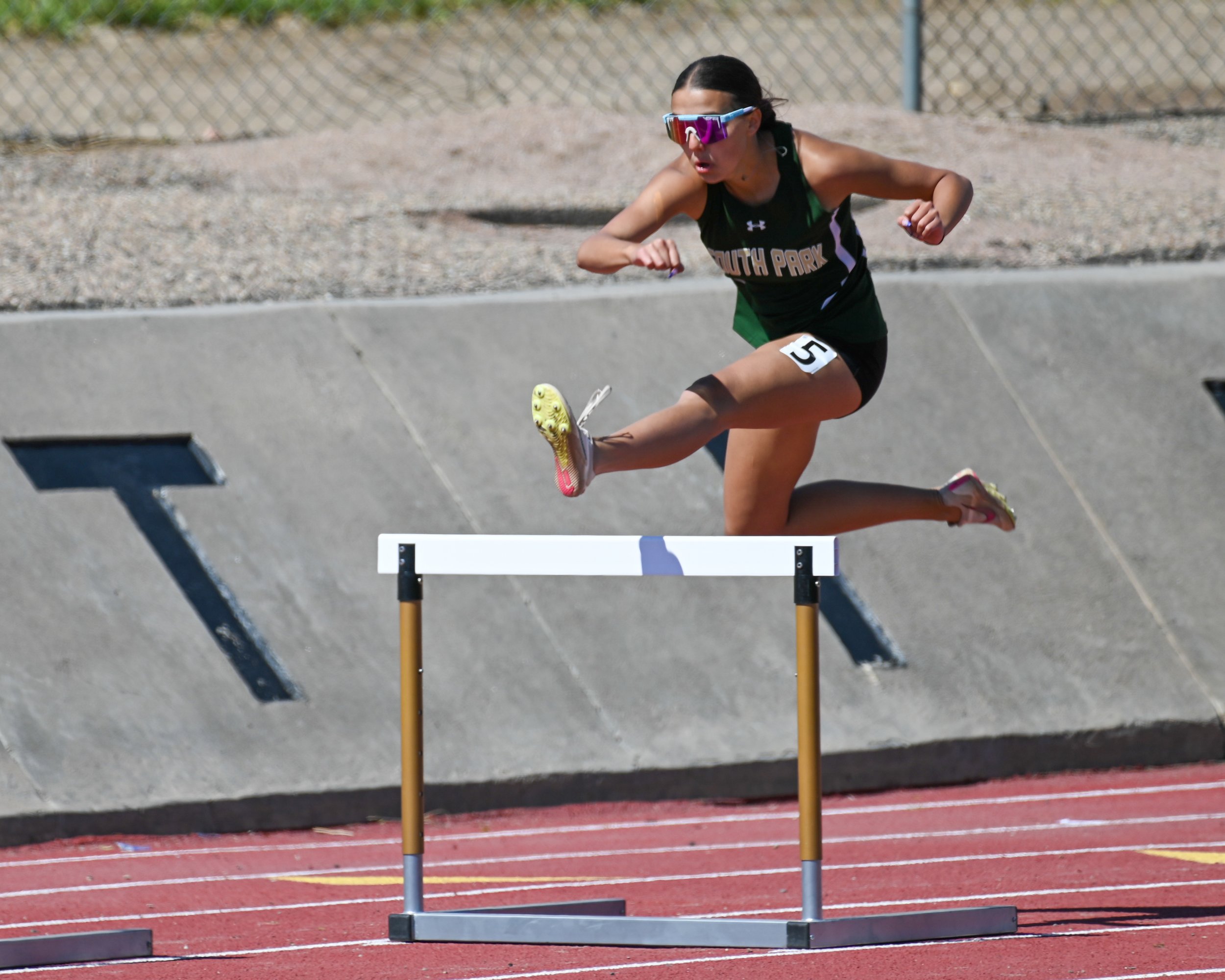 A female athlete in black athletic gear and running shoes jumps over a hurdle on a red outdoor track, wearing sunglasses. 