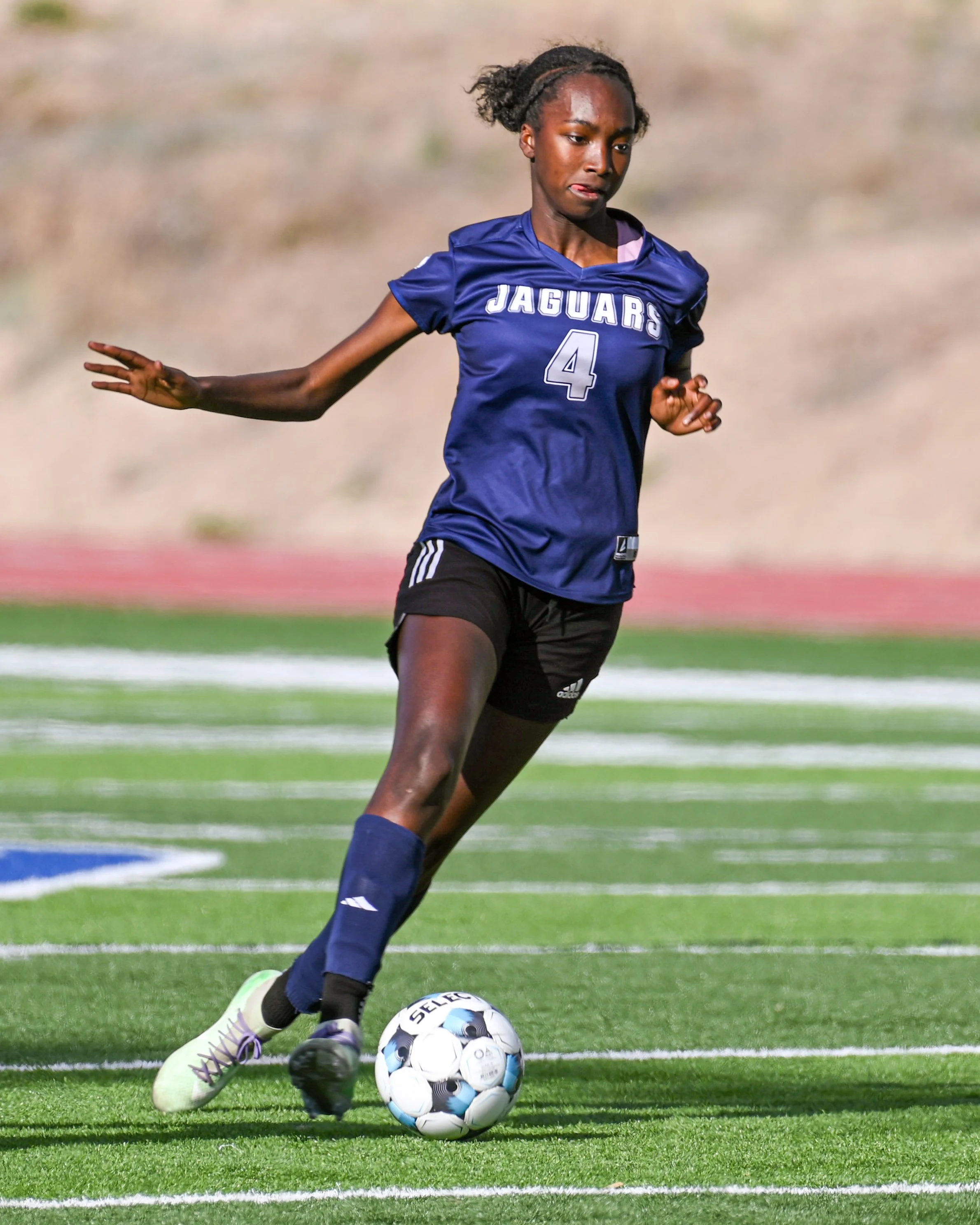 A young female soccer player wearing a blue jersey with the number 4 and 'Jaguars' on it, running on a green field, preparing to kick a soccer ball.