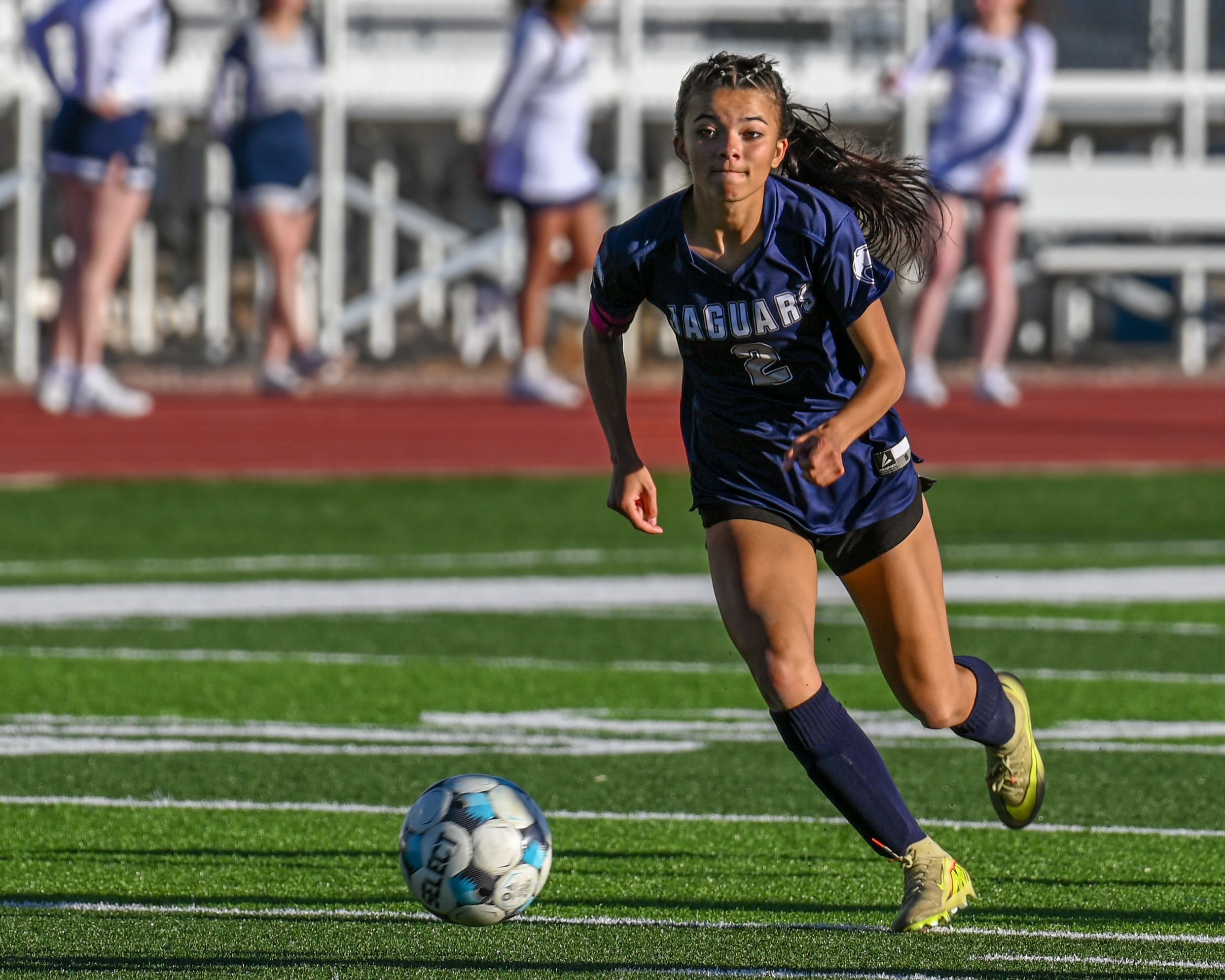 A female soccer player in a blue uniform with the number 2, running on a field and about to kick a soccer ball.