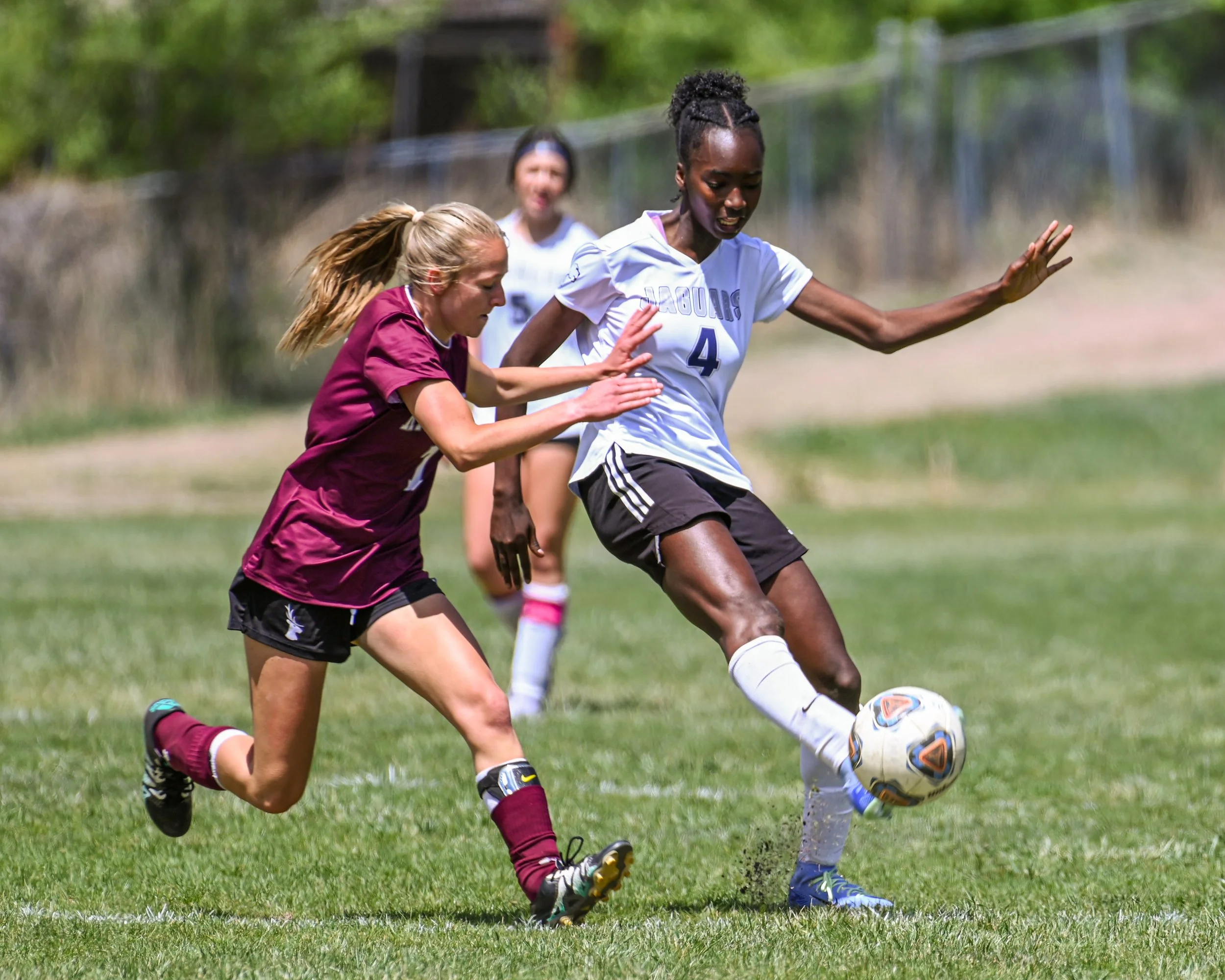 Female high school soccer players competing for the ball on a grassy field, with trees in the background.