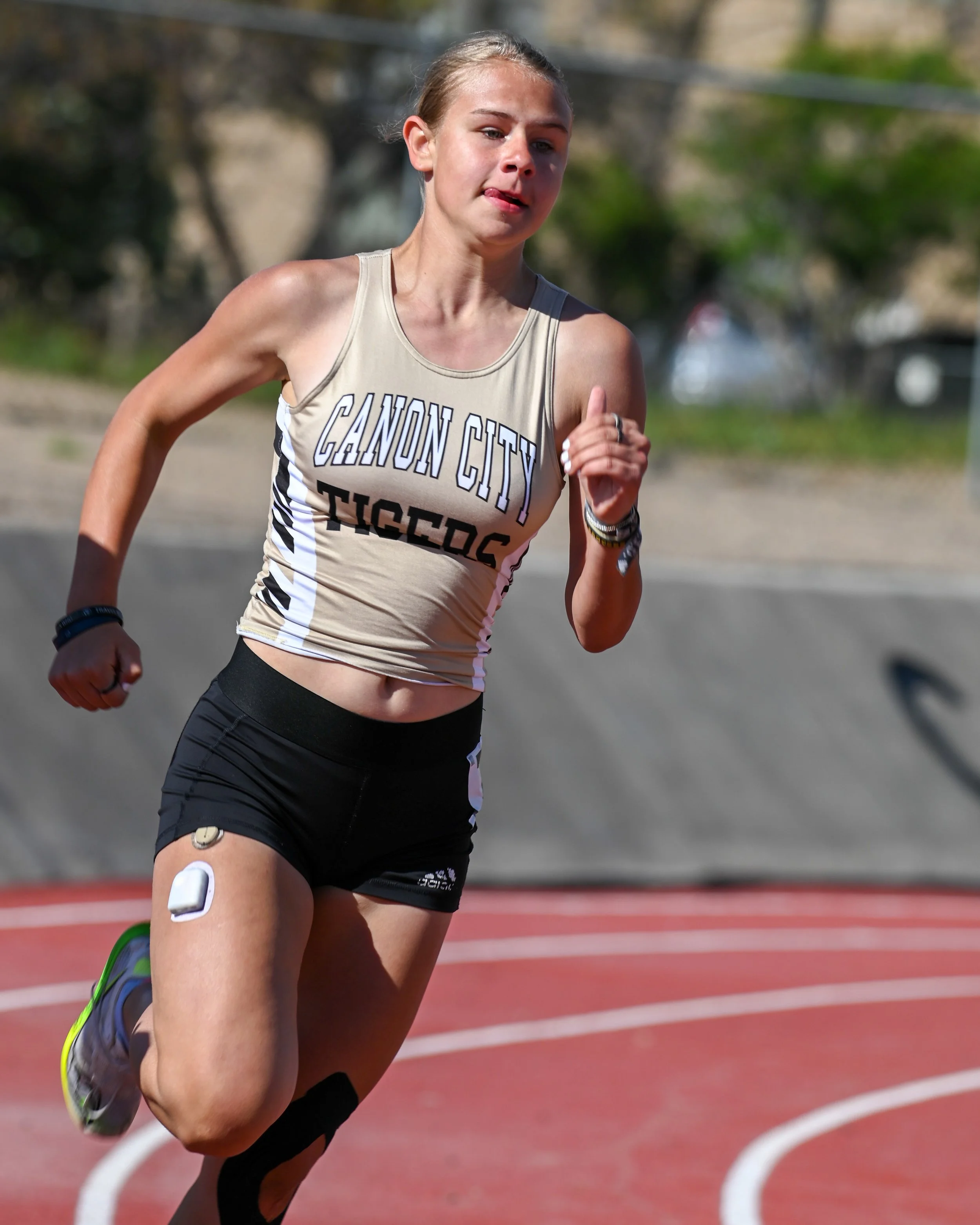 A female athlete running on a track, wearing a beige tank top that reads 'Cañon City Tigers' and black shorts. She appears focused and determined.