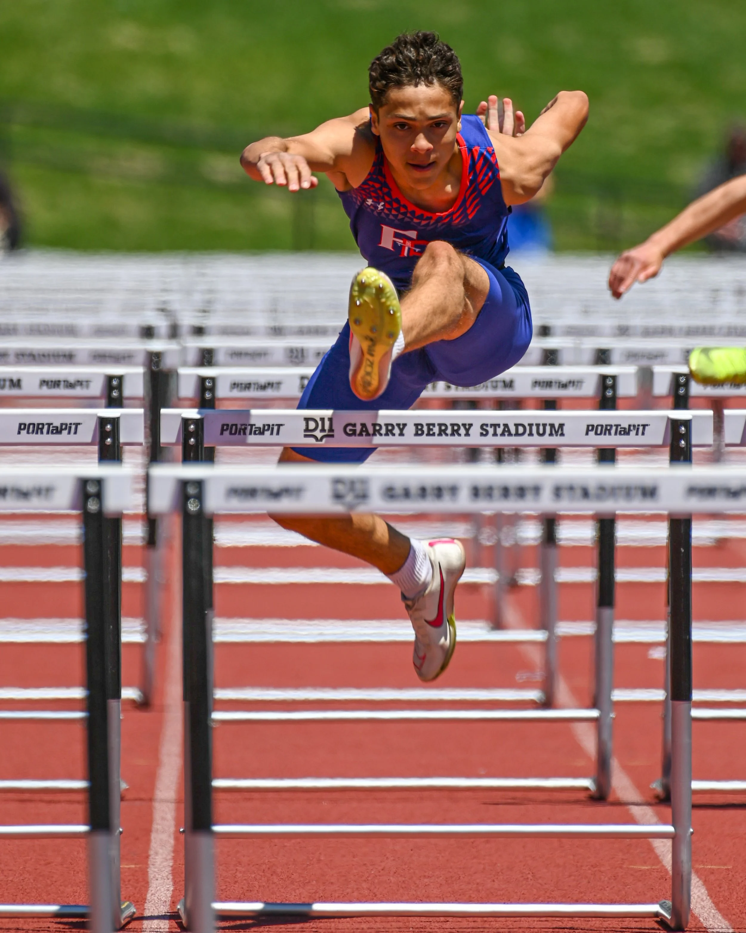 A male hurdler from Fountain Fort Carson jumping over hurdles on a red running track at Garry Berry Stadium in Colorado Springs.