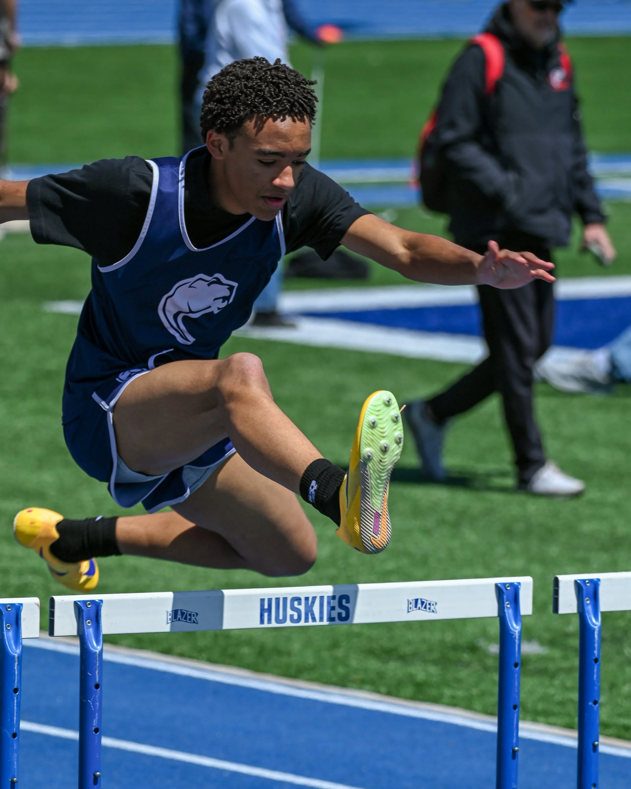 A male track athlete wearing a blue jersey is participating in a hurdle race, mid-air over a hurdle. 